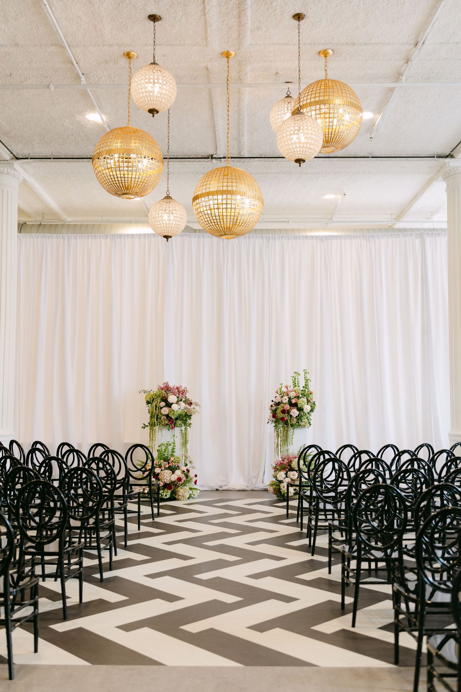 Elegant wedding ceremony venue with gold disco ball lights, black chairs, chevron floor, and floral arrangements against white draping.