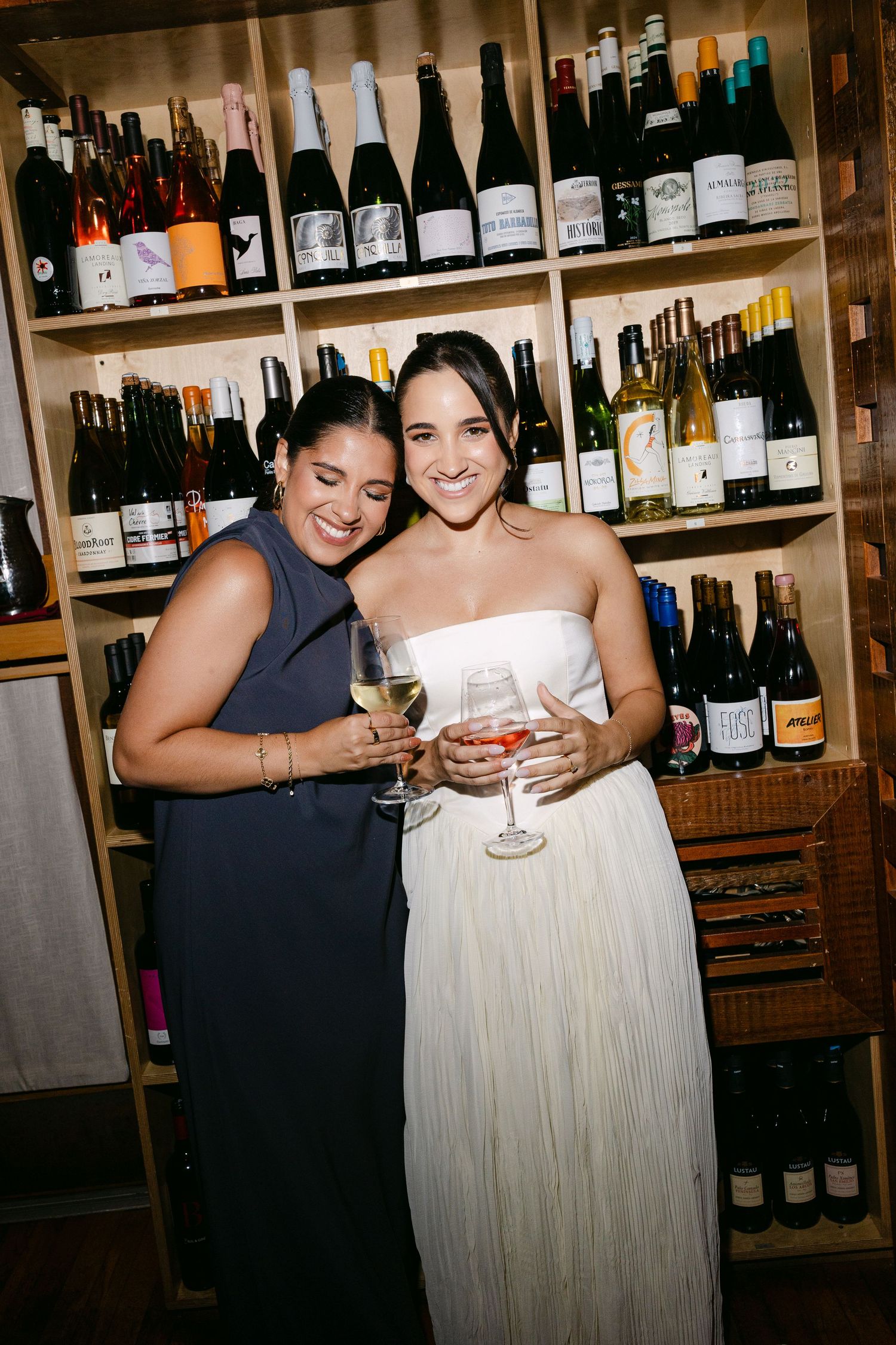 Two friends share a joyful moment together at a wine bar with bottles displayed on shelves in the background.