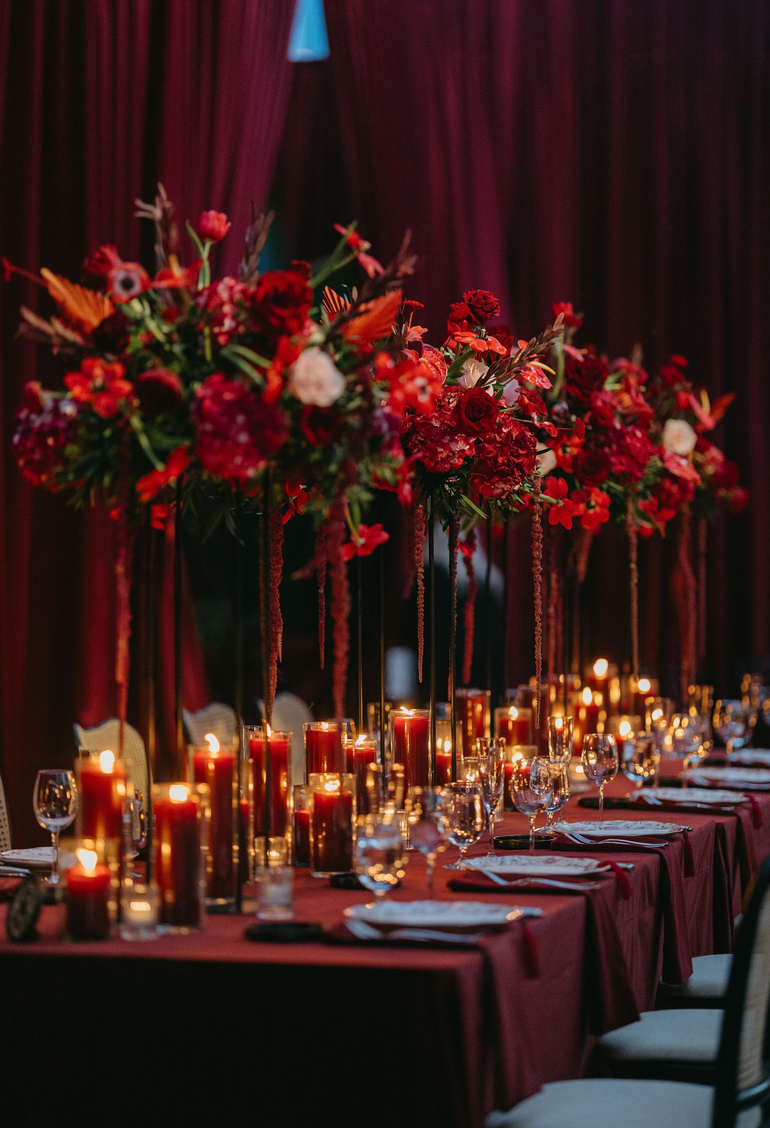 Elegant wedding reception table setup with burgundy linens and tall red floral centerpieces illuminated by glowing candlelight.