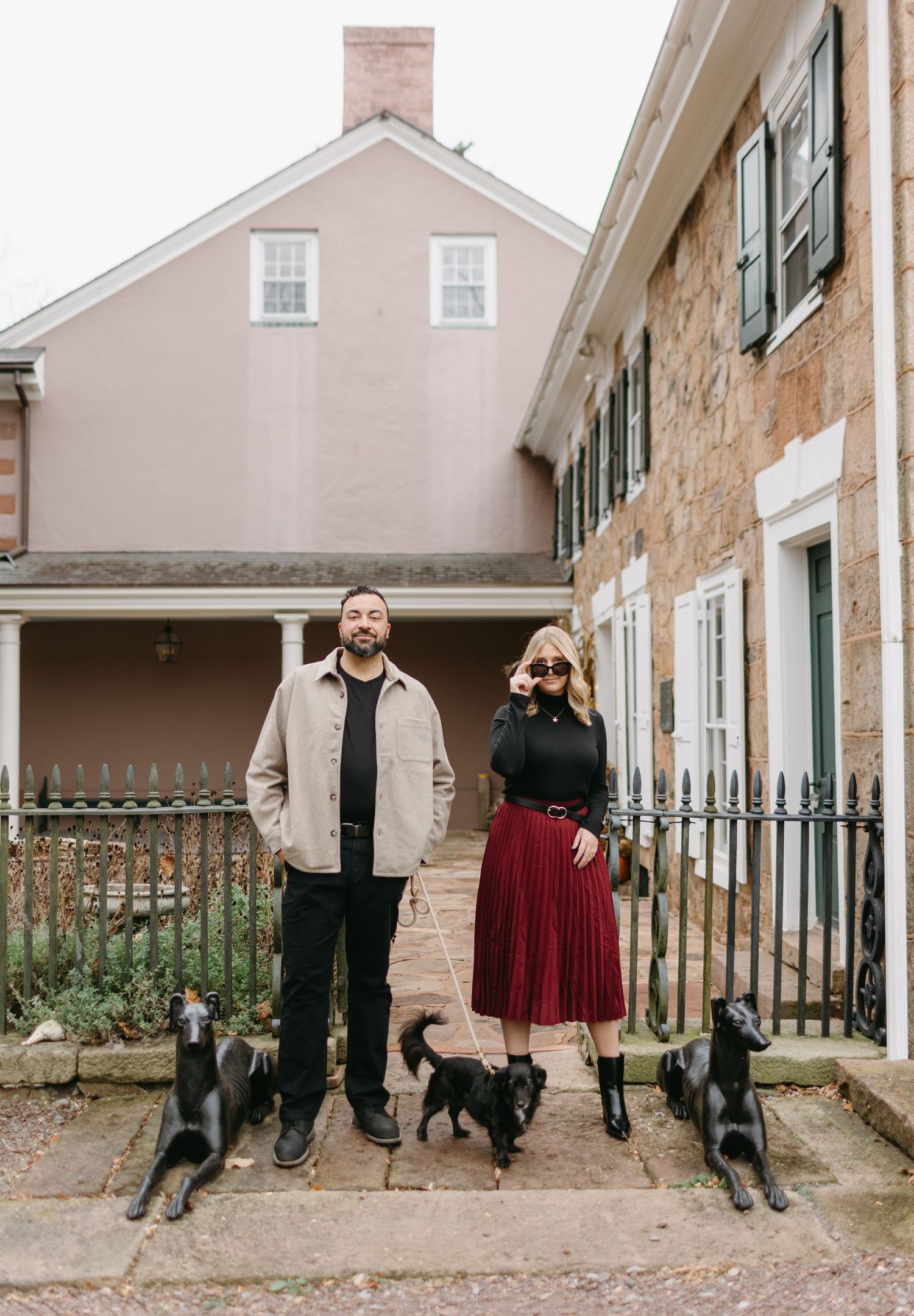A couple stands with three dogs outside a historic stone building with green shutters.