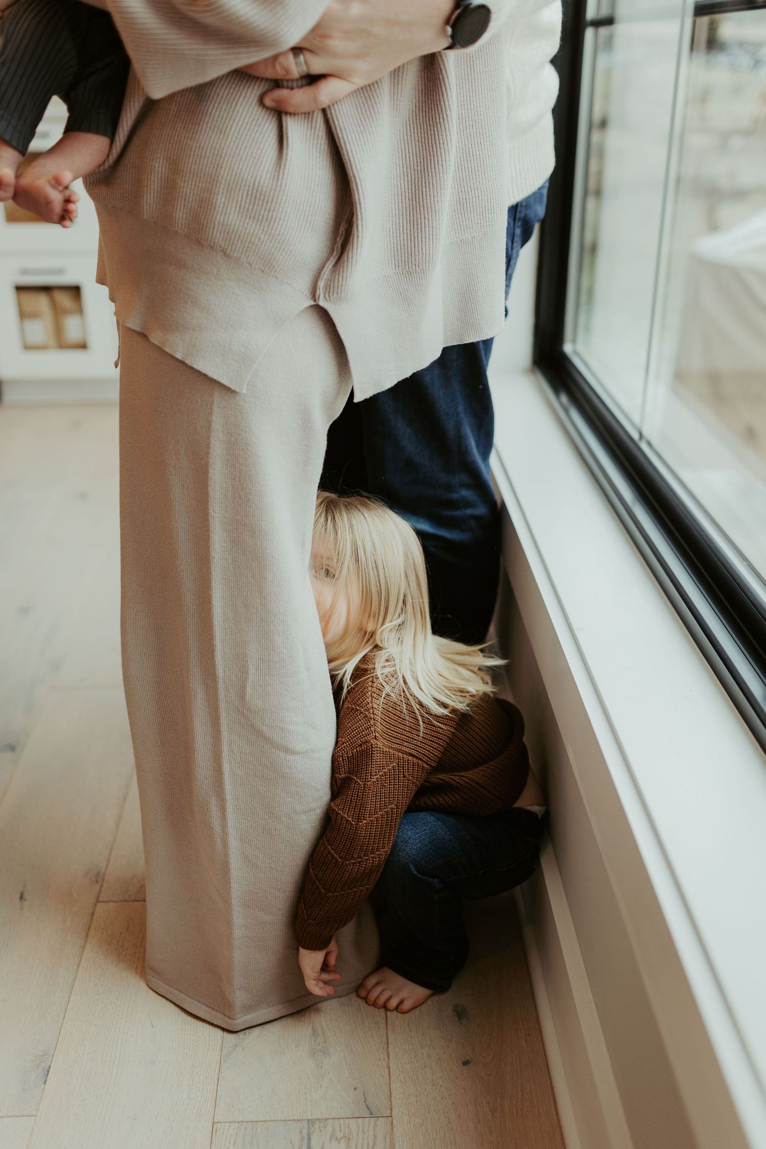 A child playfully hides behind cream-colored clothing while peeking out by a window.