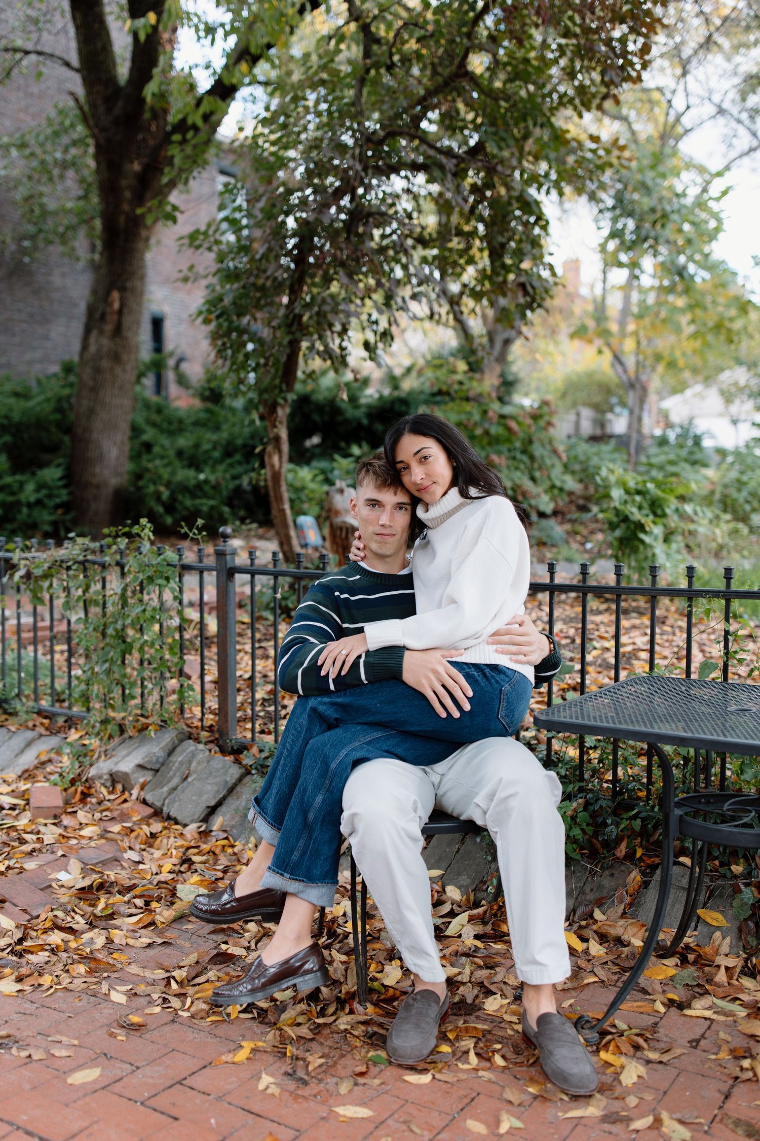 A couple relaxes on a park bench surrounded by fallen autumn leaves and iron fencing.