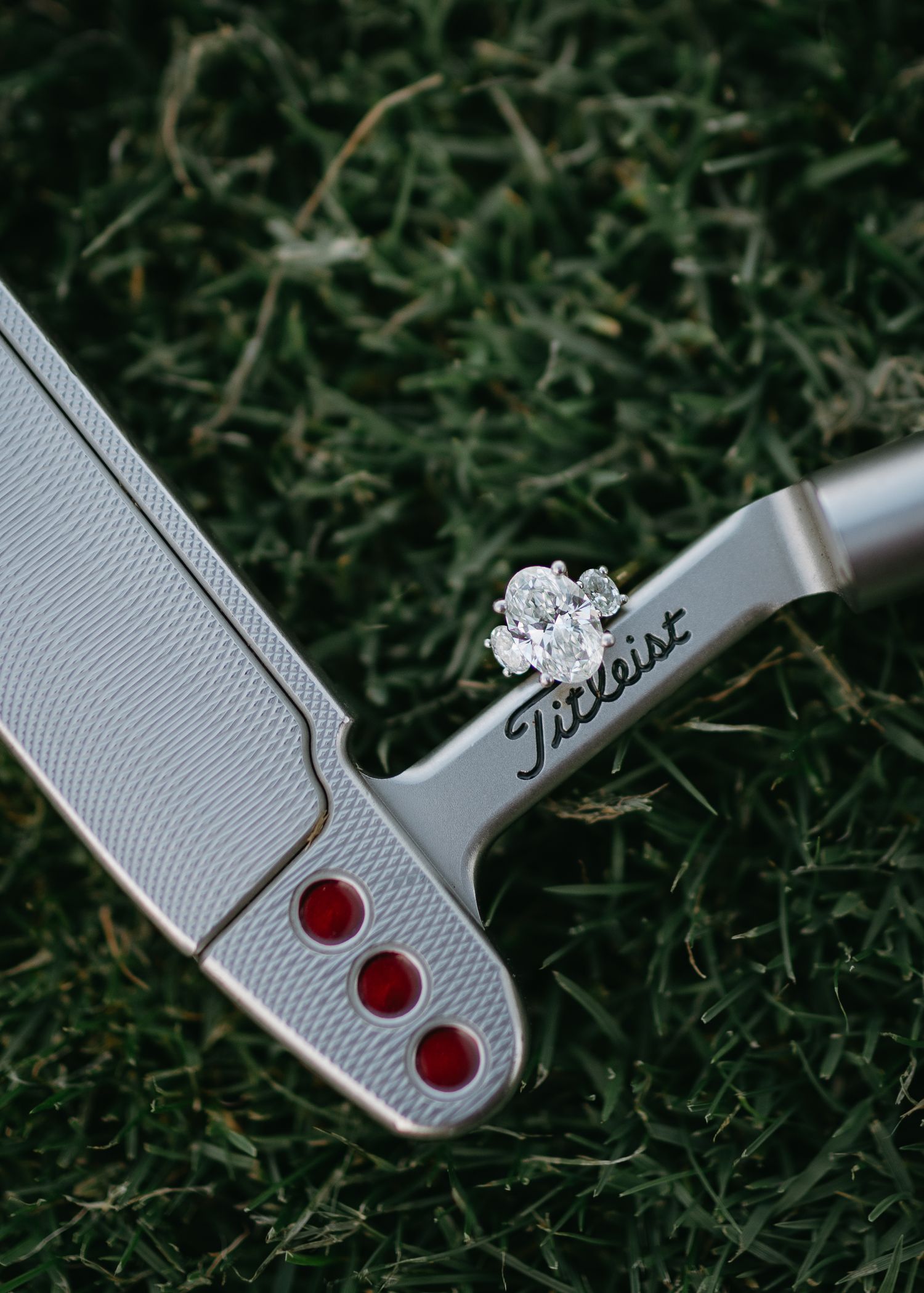 Engagement ring displayed on a Titleist golf putter against grass background.