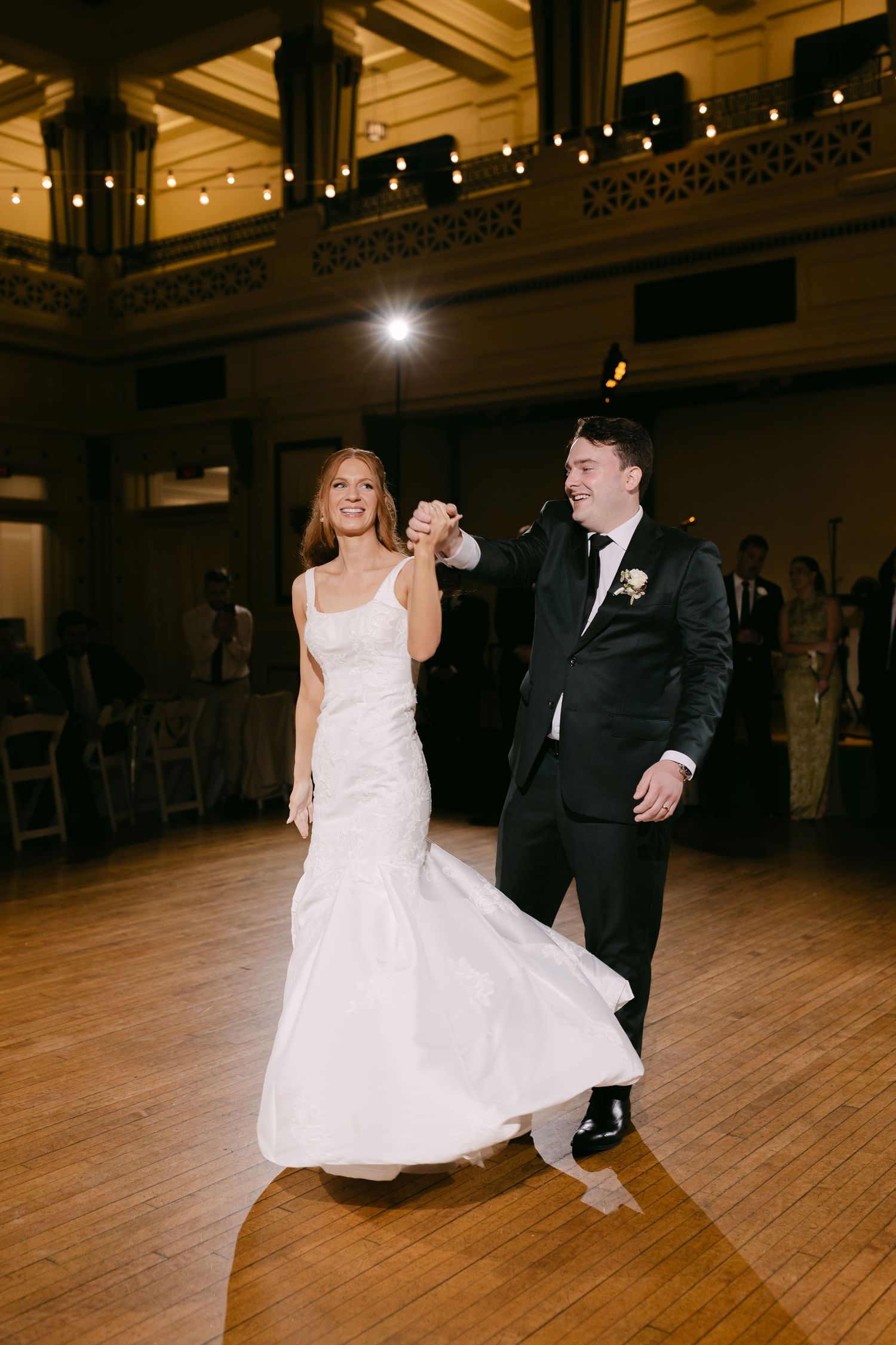 Bride and groom share their first dance, smiling joyfully in an elegant ballroom with warm string lights.