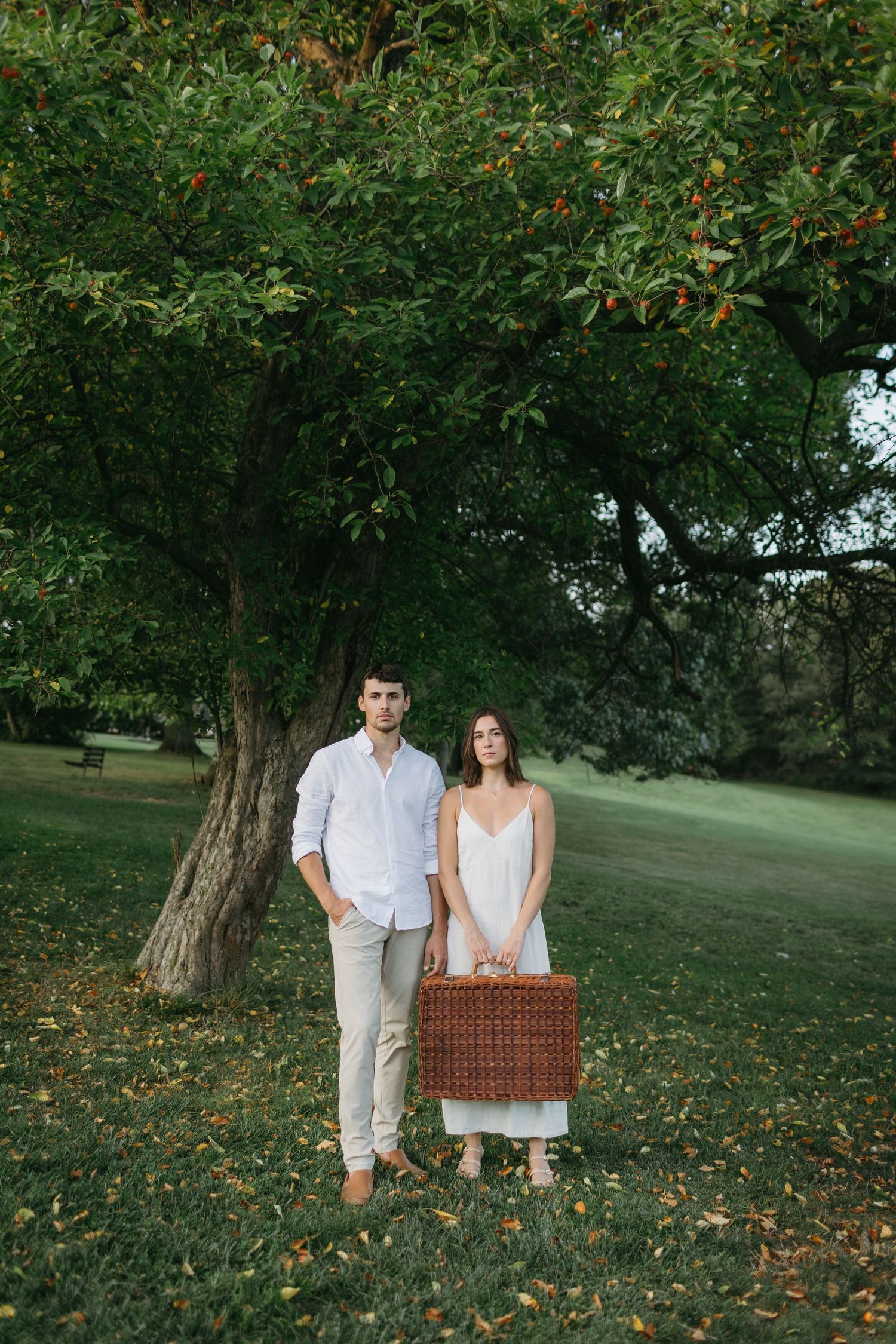 Couple in white outfits stands with picnic basket under tree in lush park setting.