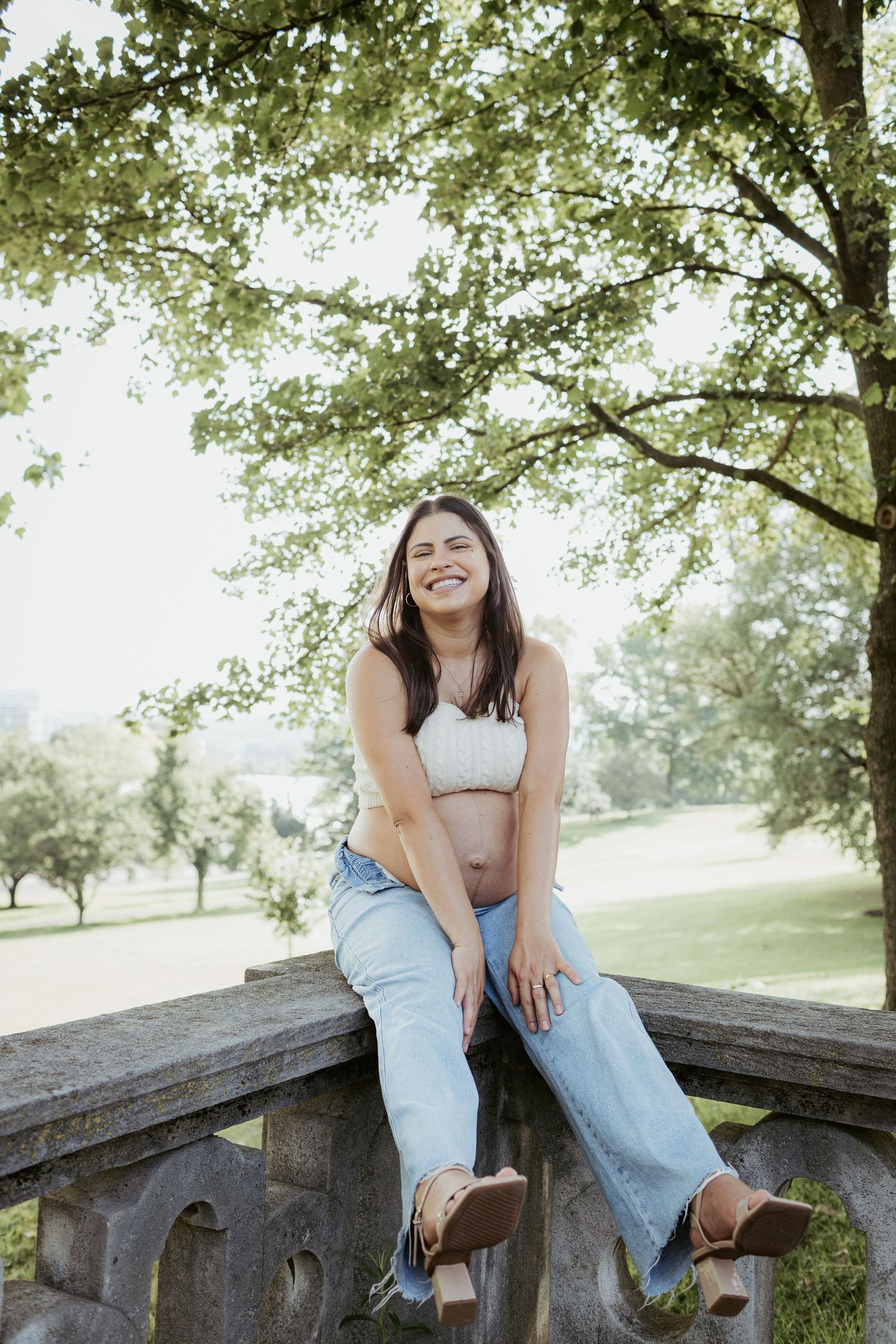 A person sits casually on stone railing wearing jeans and crop top in sunny park setting.