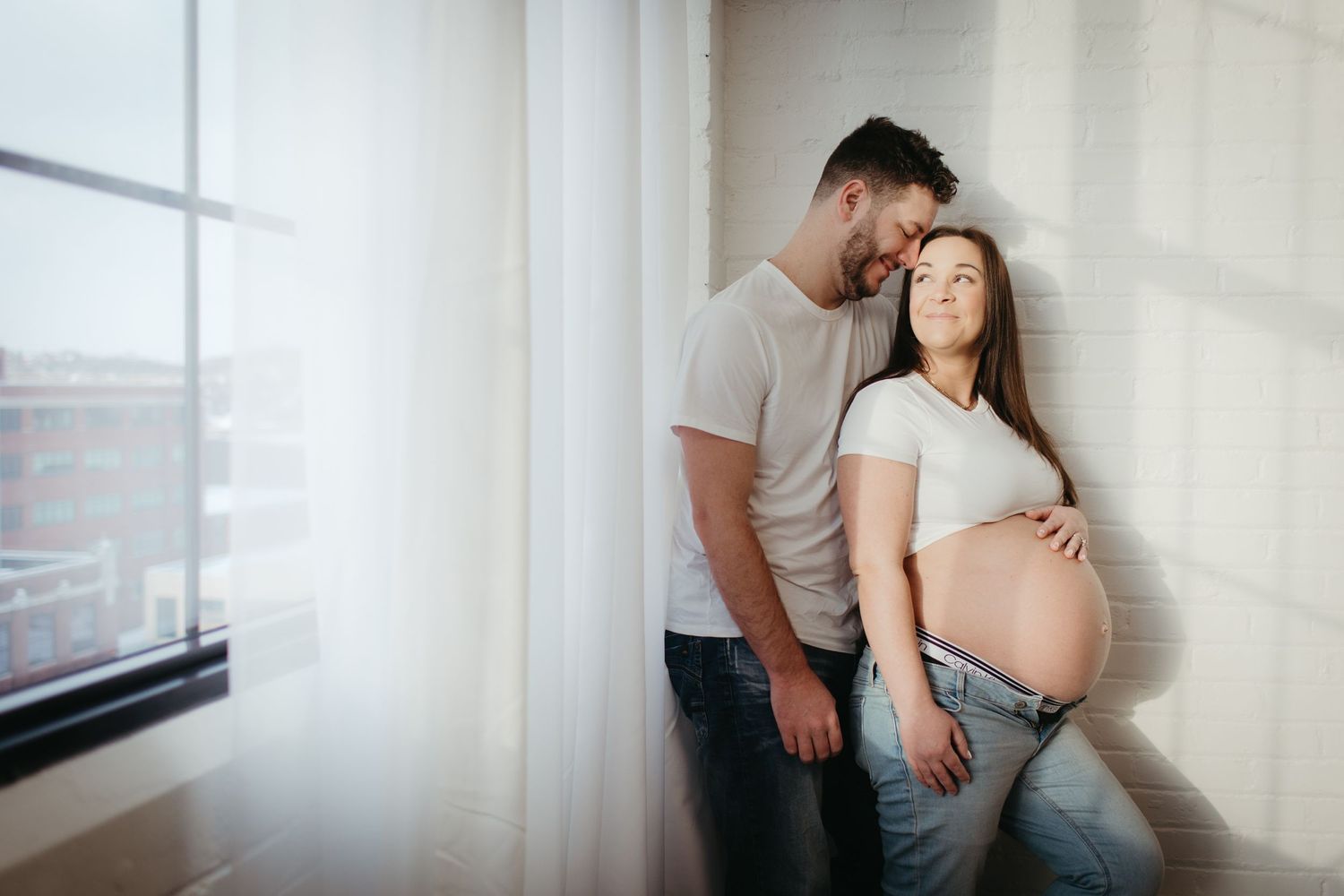 Intimate maternity photo of a couple by a window in soft natural light.
