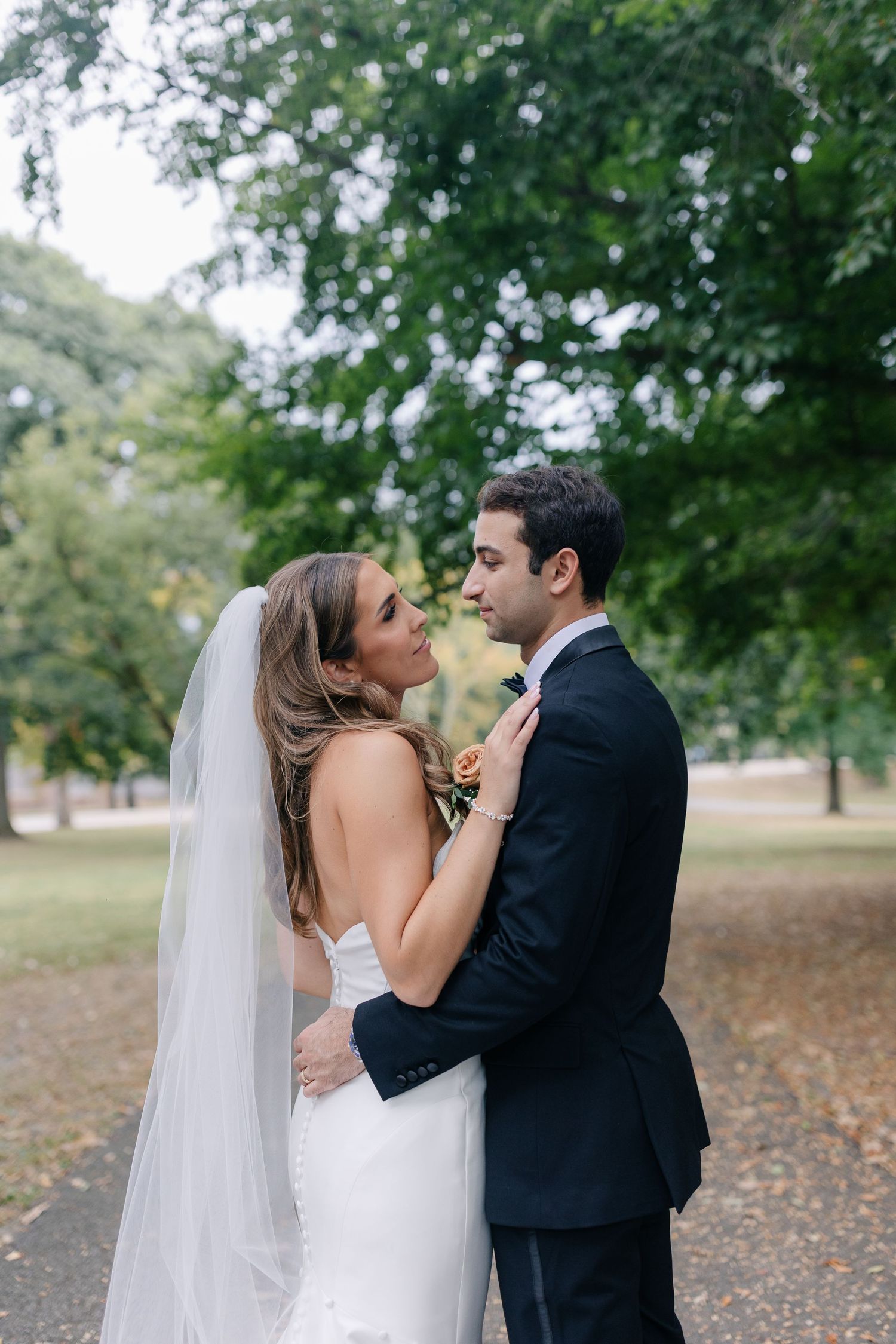 A romantic wedding portrait of a couple embracing outdoors beneath green trees on their special day.