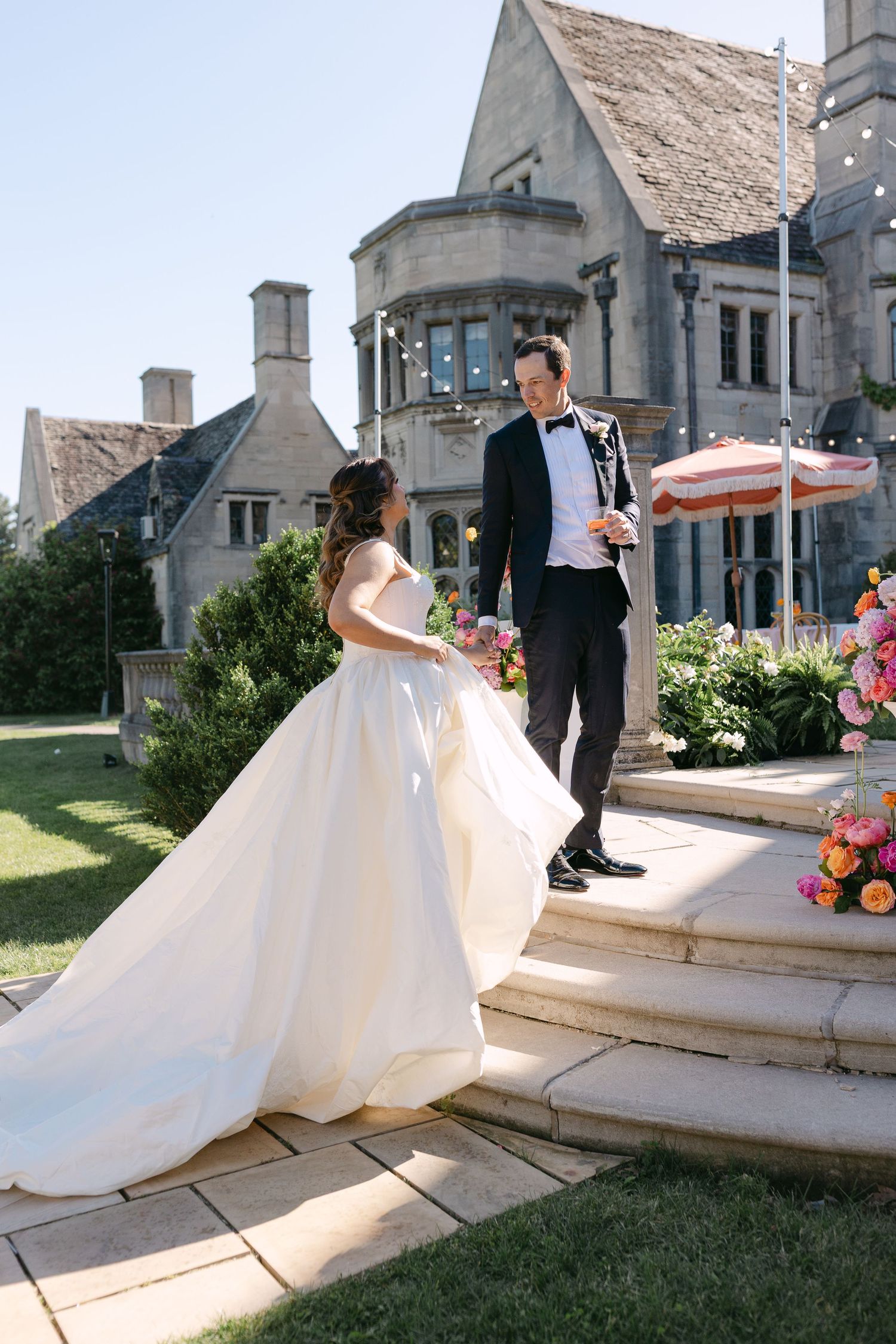 Bride in white ball gown and groom in tuxedo share a moment on stone steps outside an elegant manor.