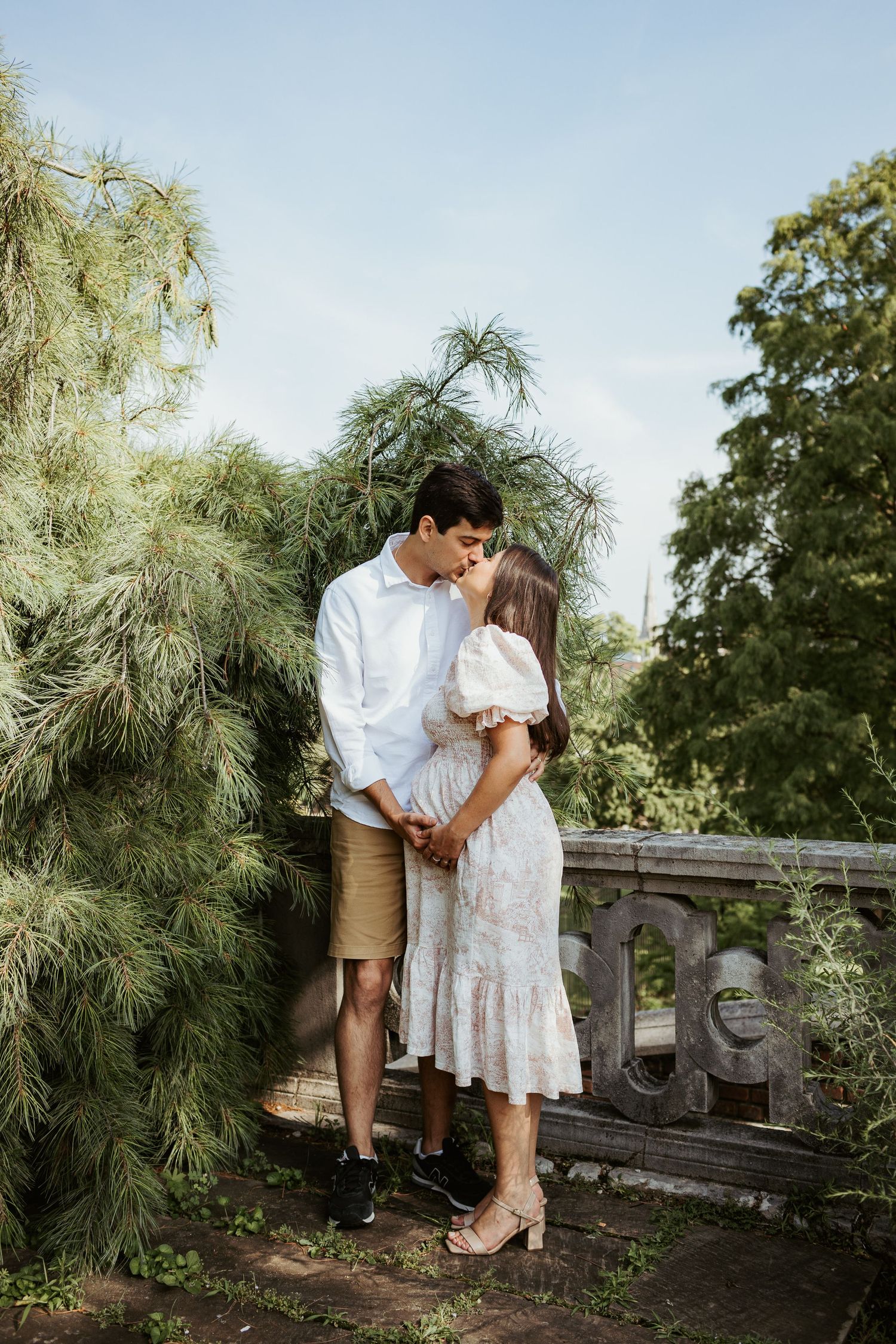A romantic couple embraces near pine trees and stone railing in outdoor setting.