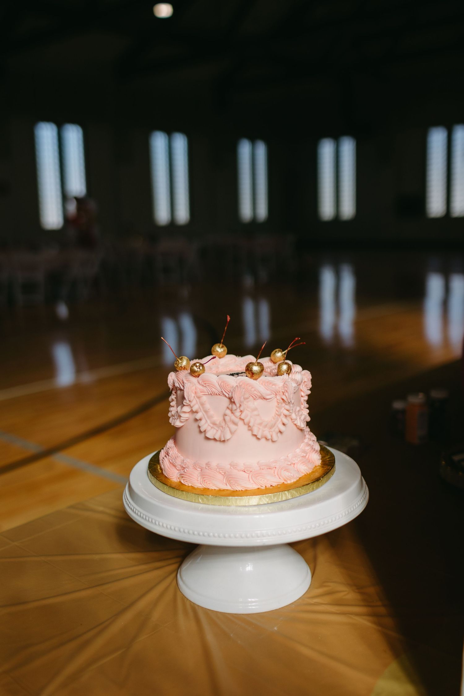 A delicate pink frosted cake displayed on a white pedestal stand in a dim room.