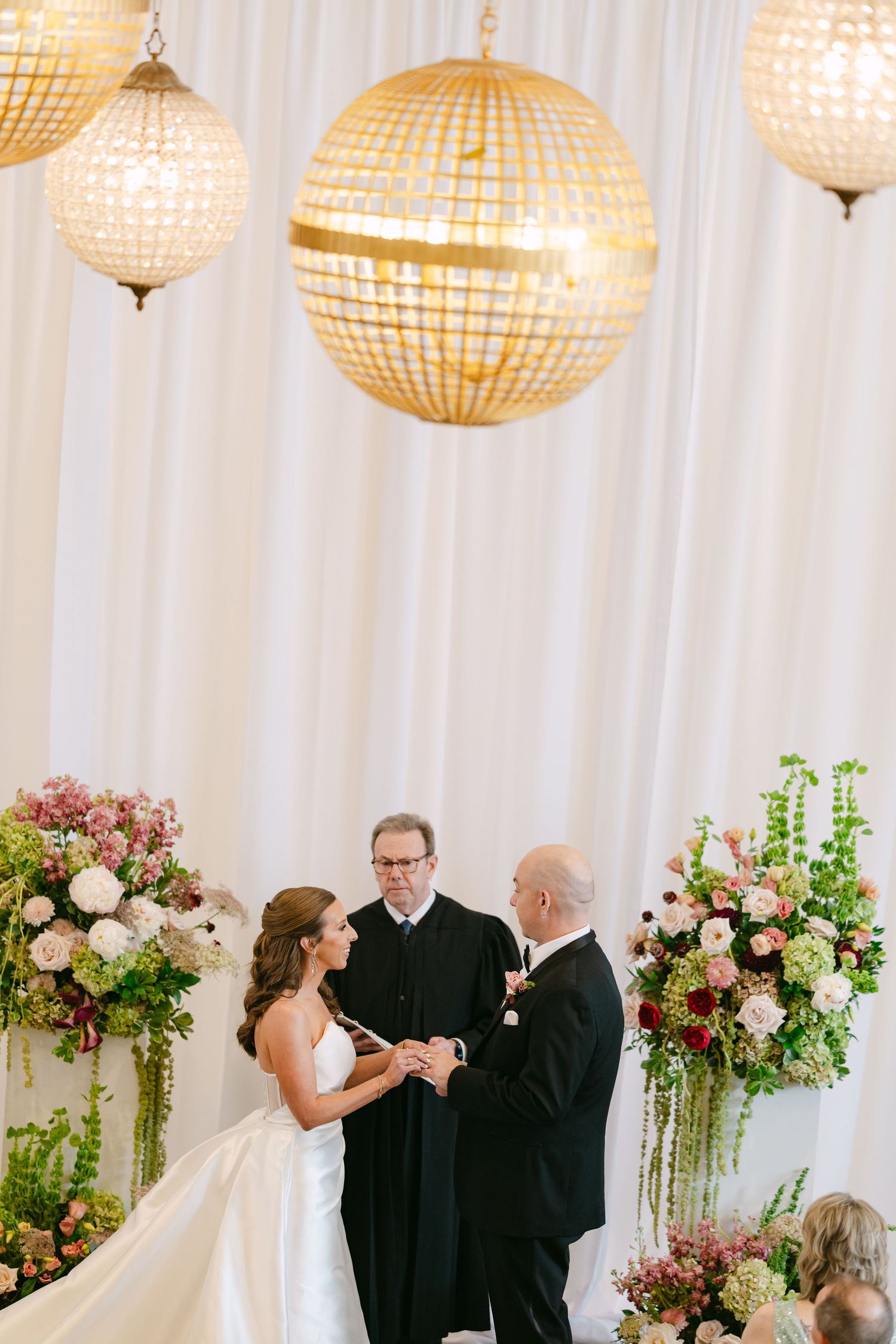 Bride and groom exchange vows beneath gold globe chandeliers, flanked by lush floral arrangements, with officiant at white draped altar.