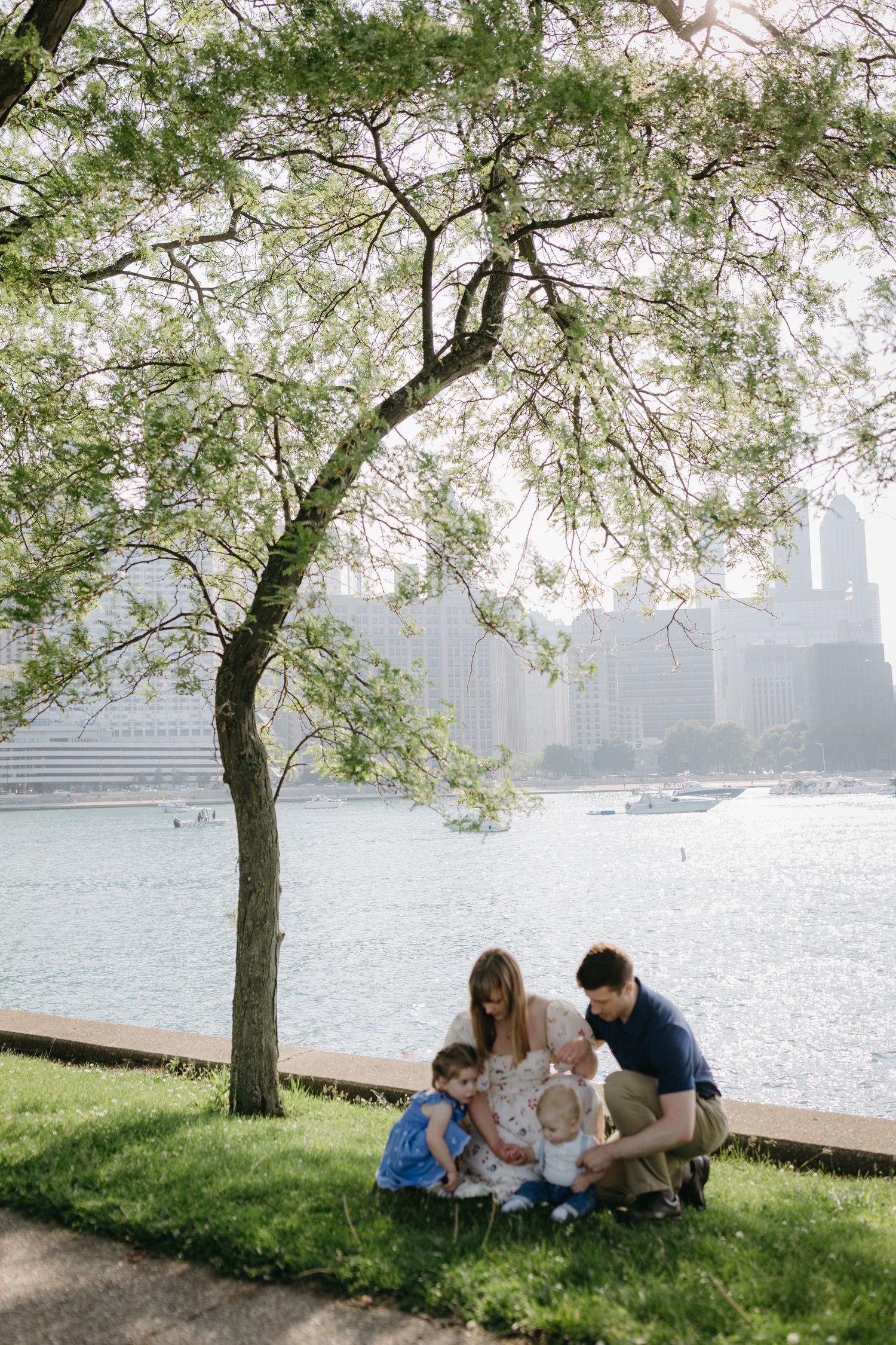 Family enjoying springtime under a blossoming tree by the water with city skyline in the misty background.