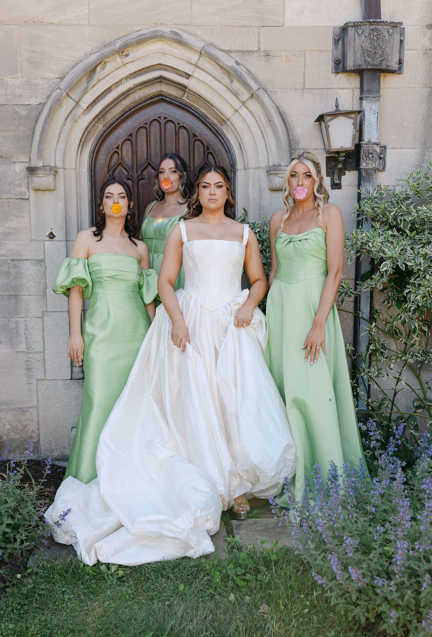 Bride in white gown poses with three bridesmaids in sage green dresses, blowing bubble gum outside a stone chapel.