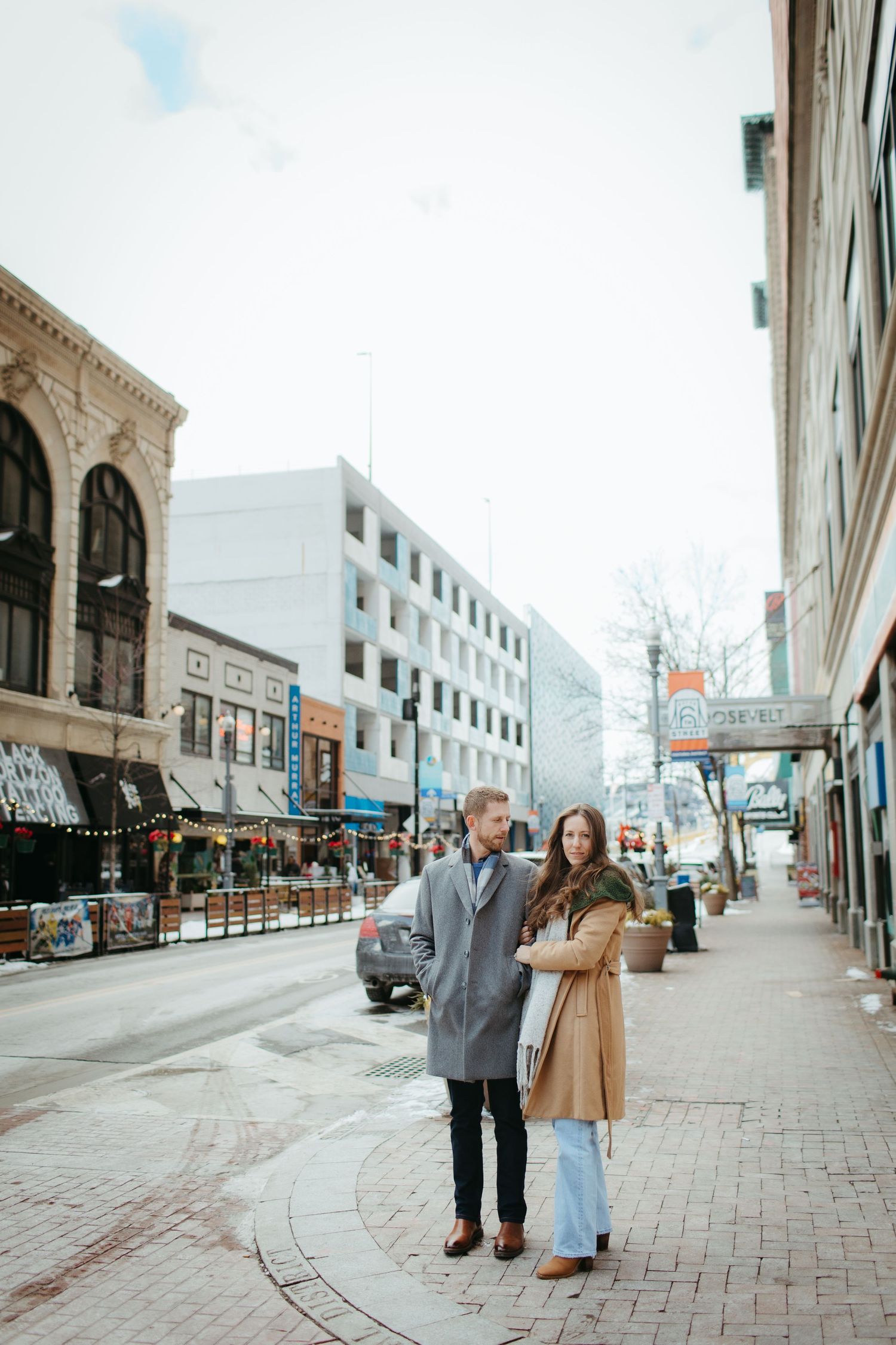 A couple stands together on a brick-paved city street surrounded by historic buildings.