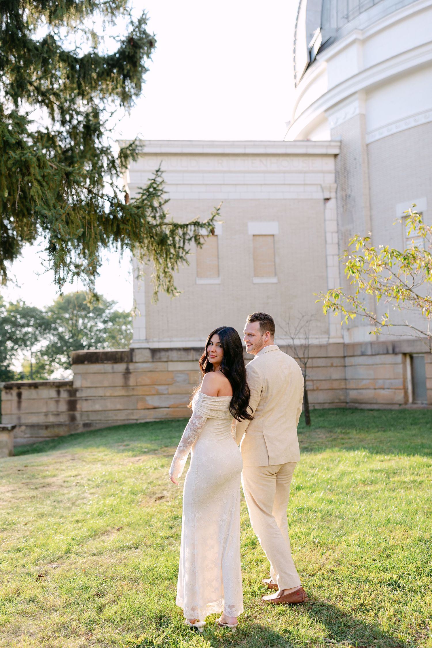 A couple in light-colored outfits shares an intimate moment on a grassy hill near a white stone building at sunset.