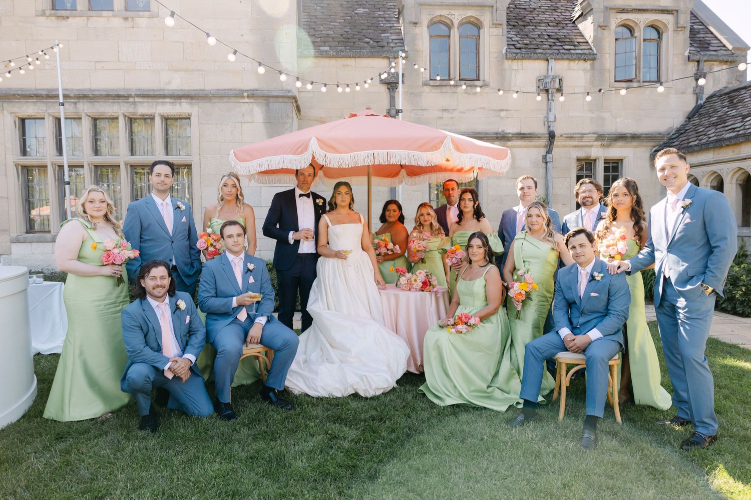 Wedding party poses outdoors at elegant estate with bride, groom, groomsmen in blue suits, and bridesmaids in green dresses.