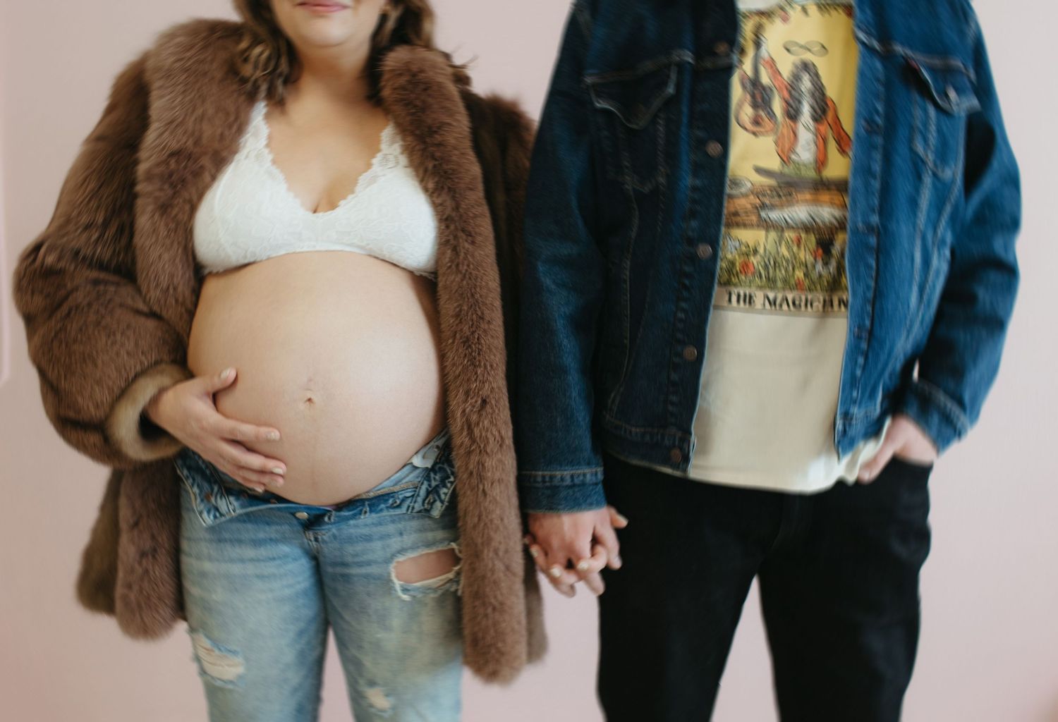 A couple holds hands, showing a pregnant belly in a white top and jeans with a brown coat and denim jacket.