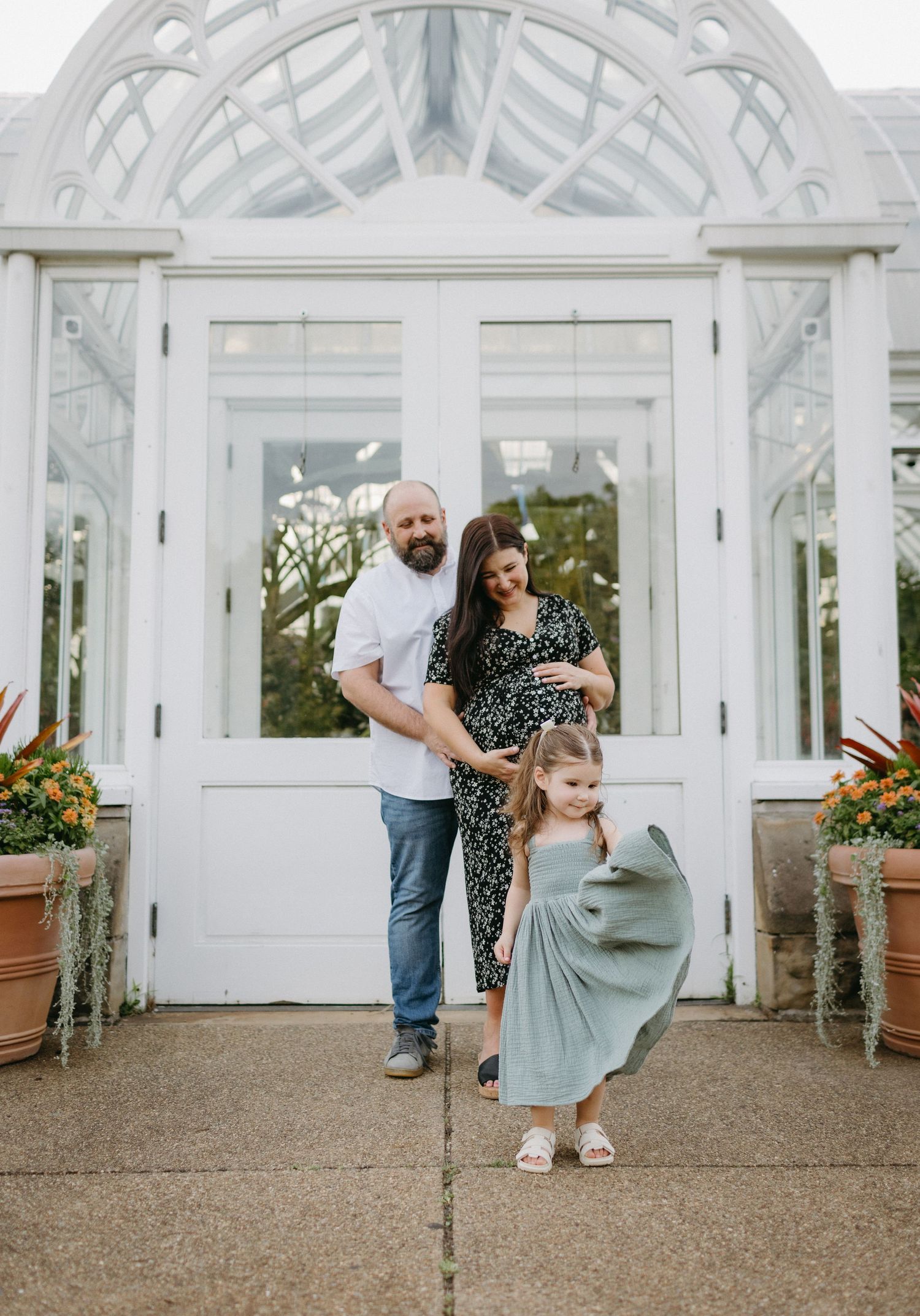 A happy family poses together in front of a white greenhouse with arched windows and potted flowers.