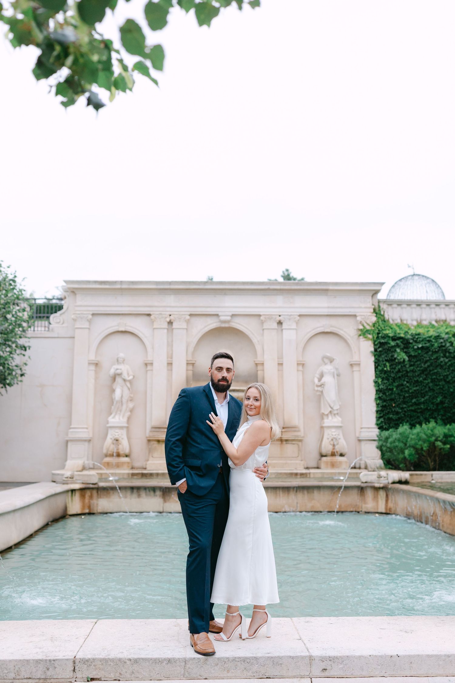Couple posing in front of an elegant fountain with classical statues, man in navy suit and woman in white dress.