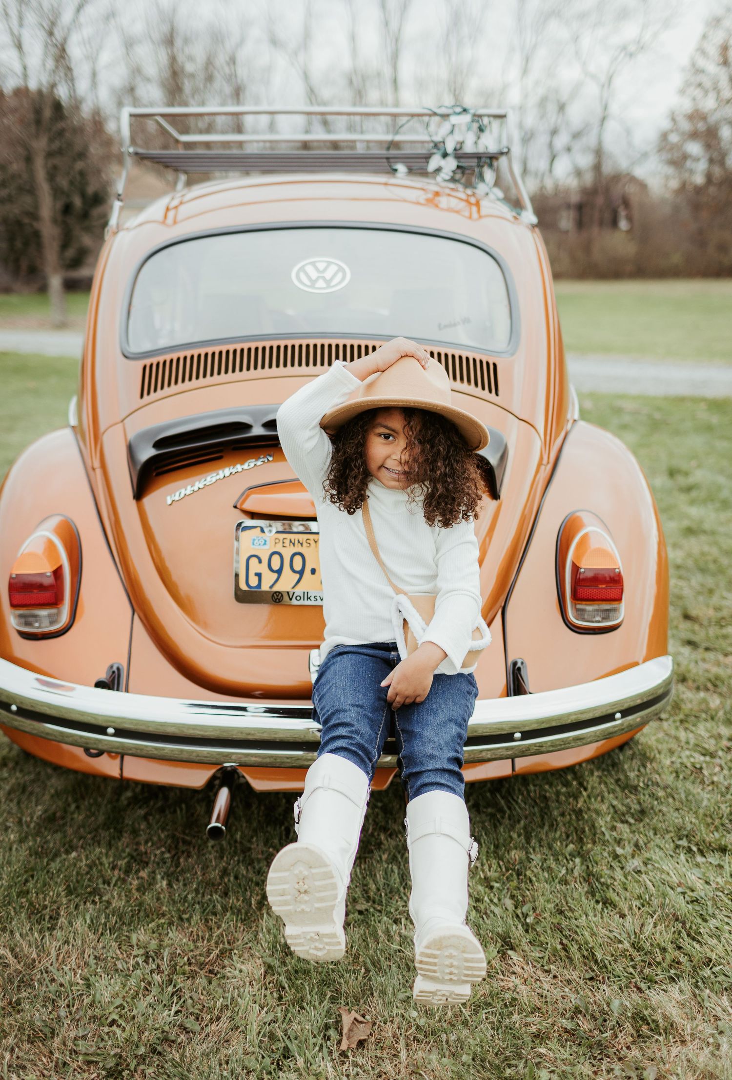 A person in white boots and denim sits on the trunk of an orange Volkswagen Beetle in a grassy field.