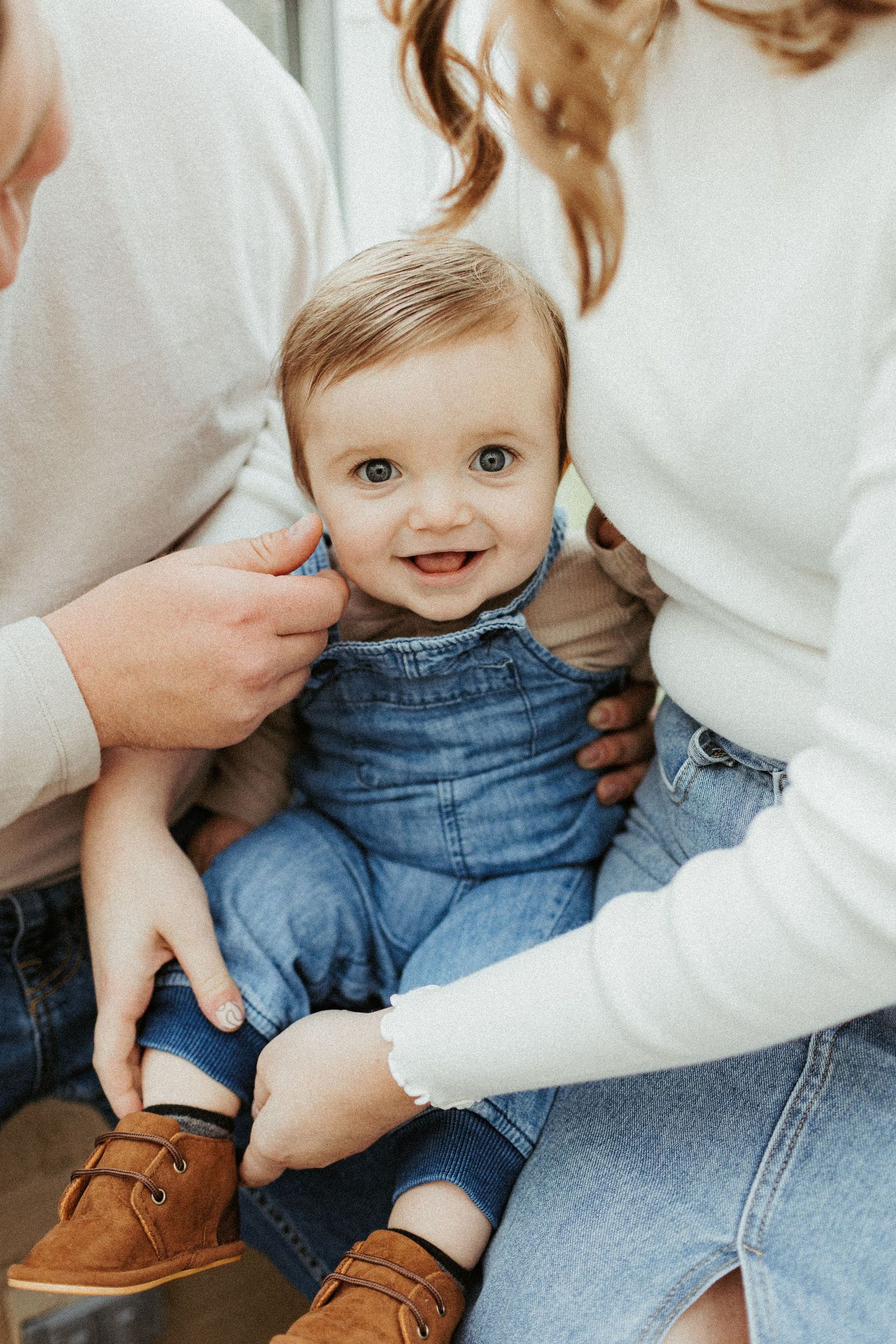Small child wearing denim overalls and brown shoes sitting between two people in white sweaters.