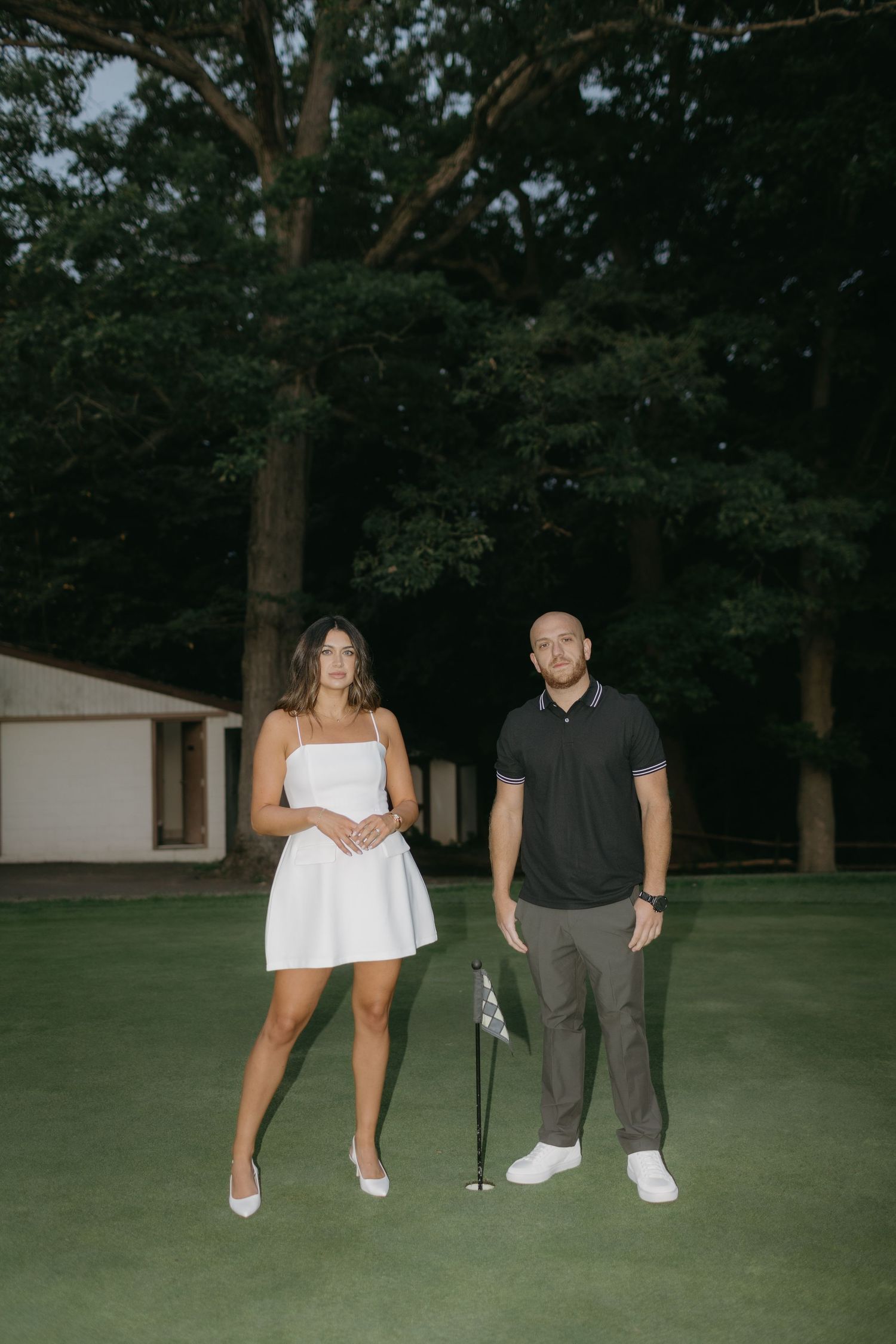 Couple standing on golf green with clubs during twilight engagement photo session.