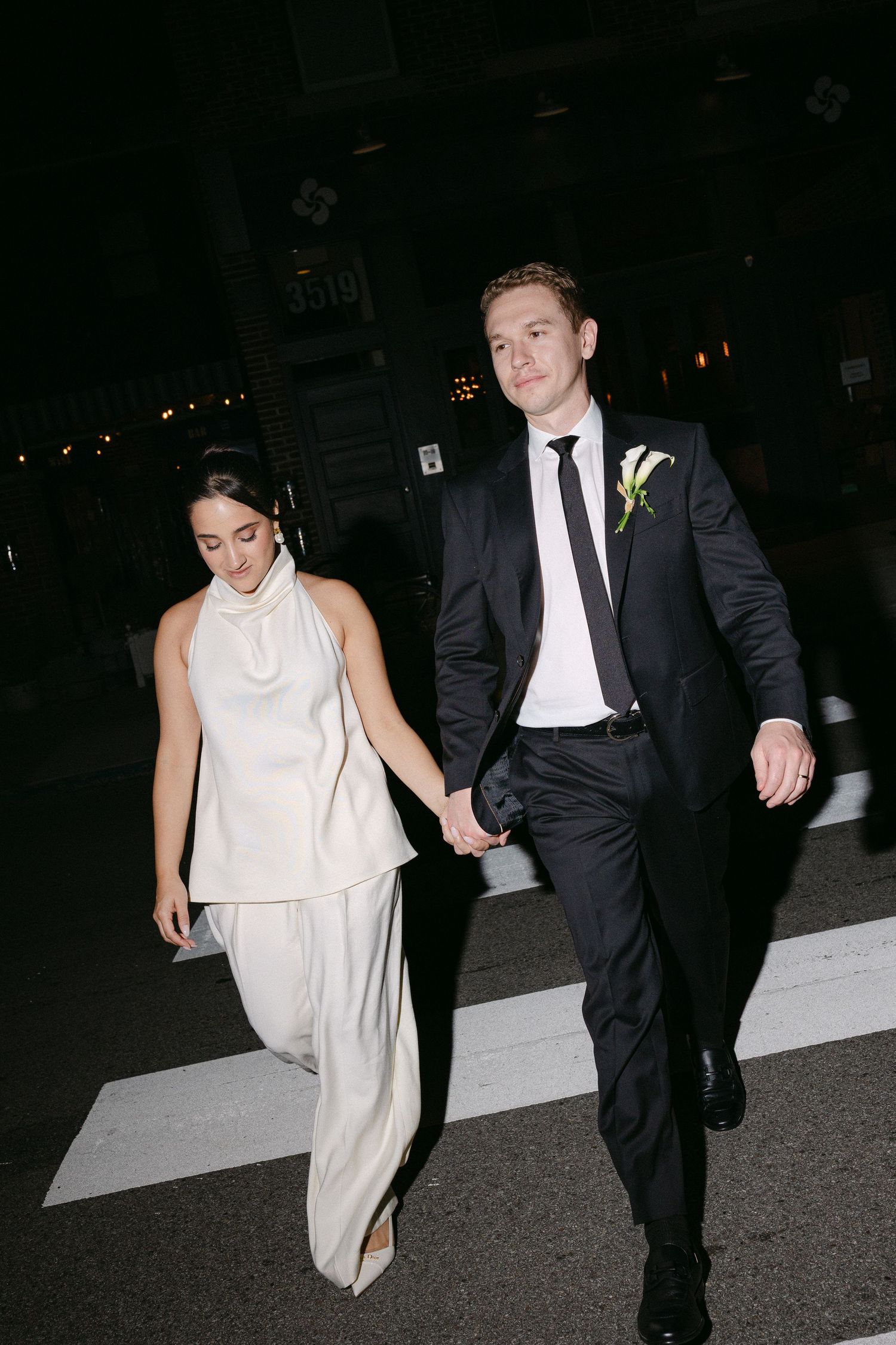 A couple in formal attire, with a woman in white dress and man in black suit, walking together at night.