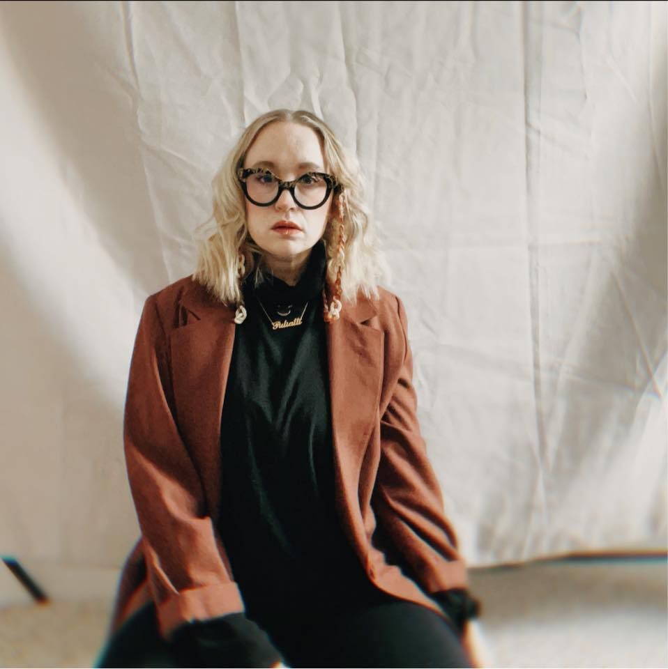 Person in a brown blazer and black turtleneck sits against white backdrop in fashion photoshoot series.