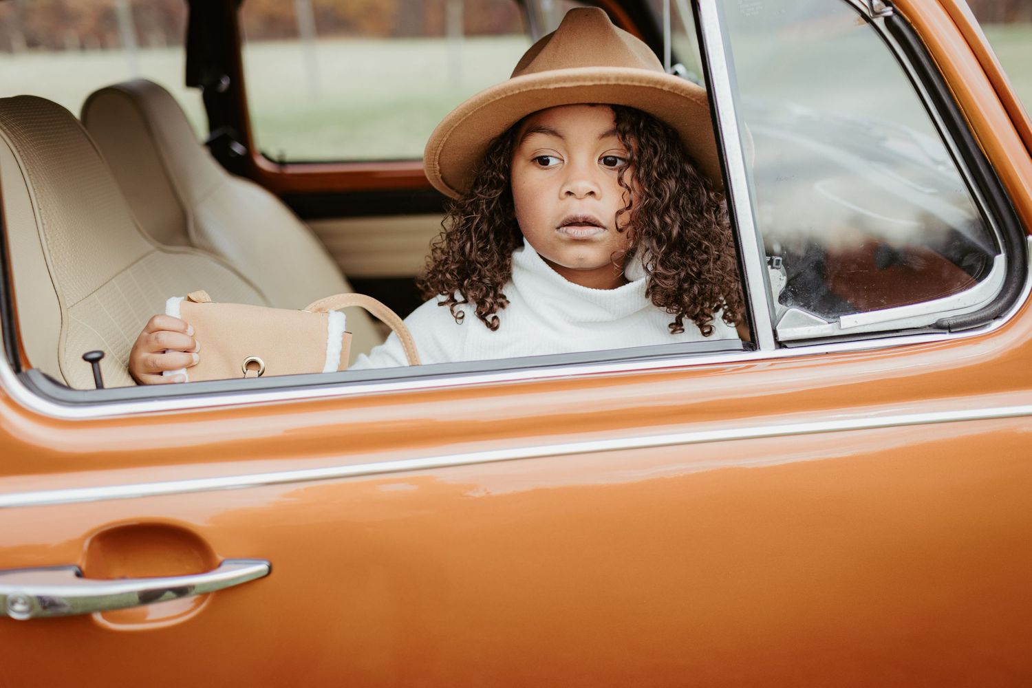 A person wearing a brown hat looks thoughtfully out the window of a vintage orange car.