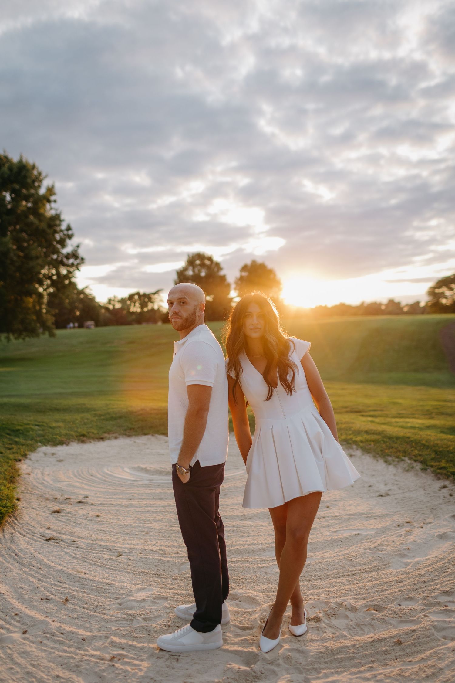 Couple in contrasting outfits pose back-to-back on sandy golf cart path at sunset.