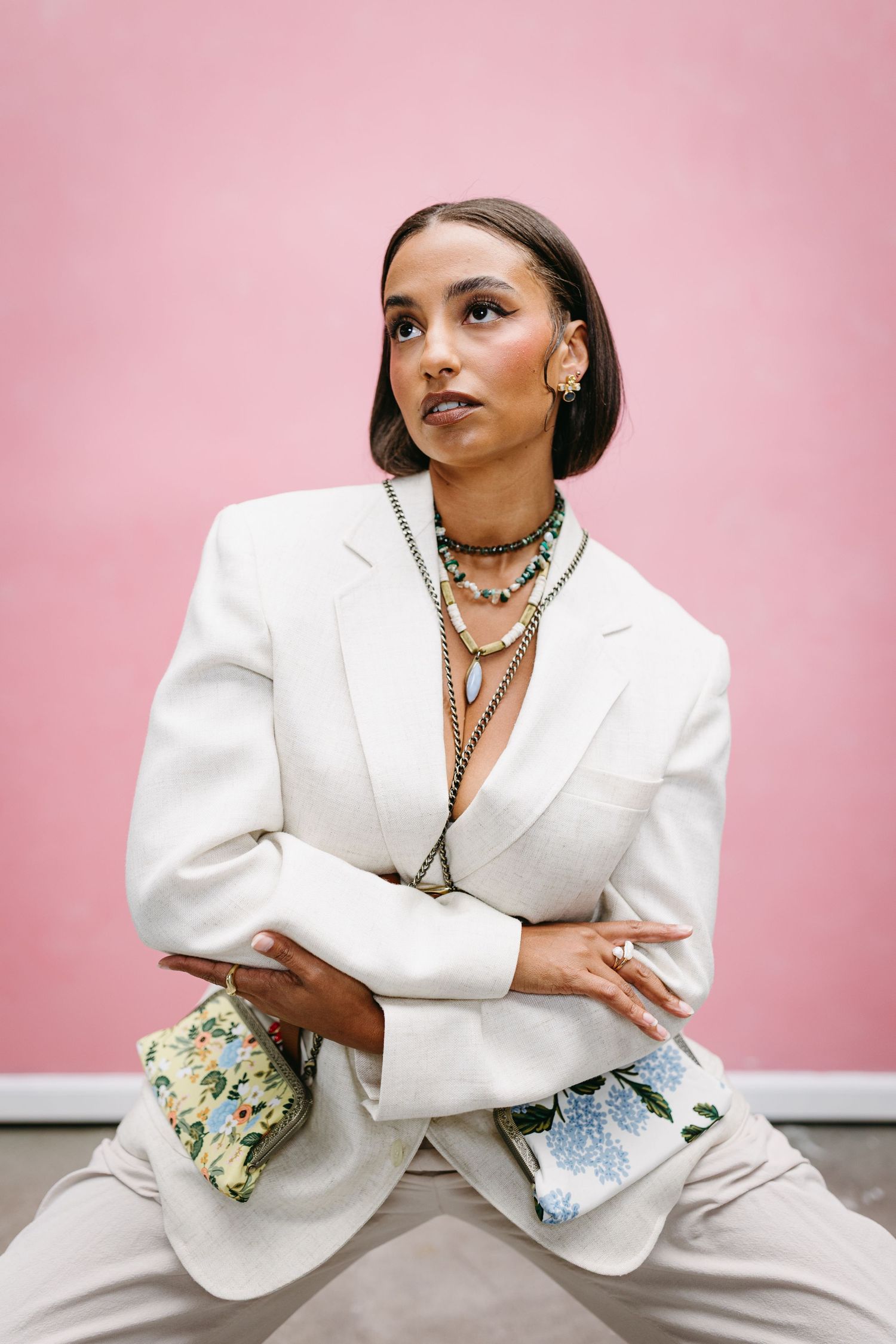 Fashionable portrait against pink background wearing white blazer, layered necklaces and neutral colored pants.