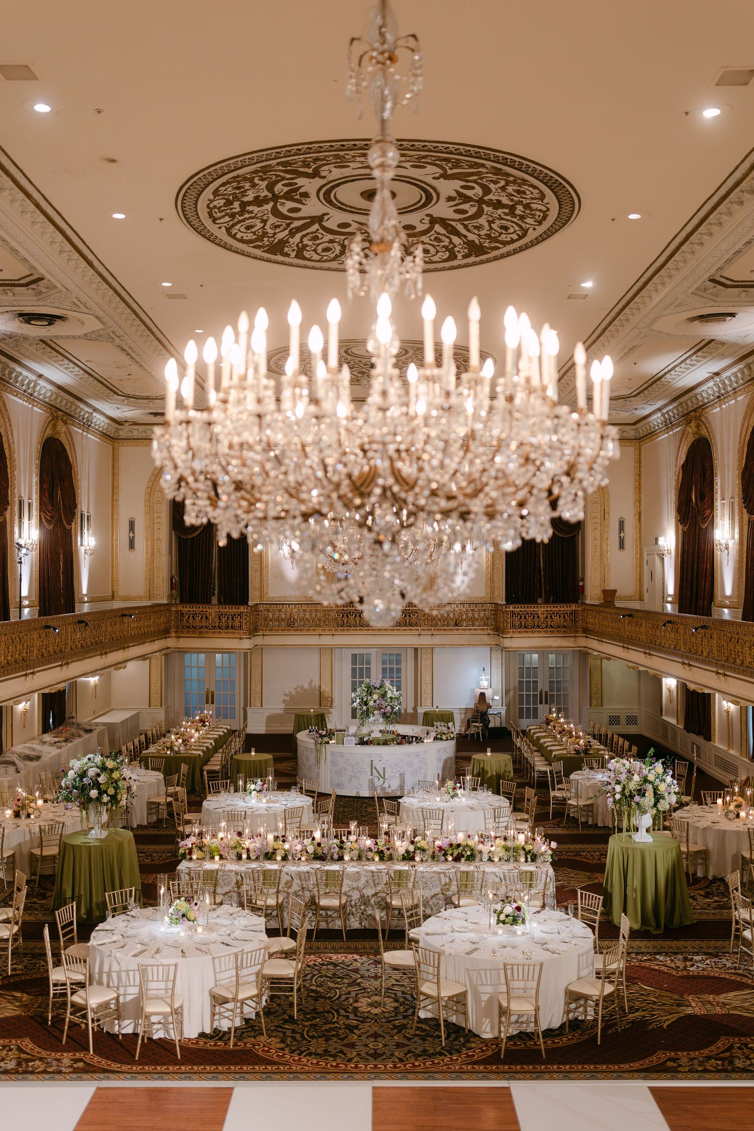 Grand ballroom wedding reception setup with crystal chandelier and round tables decorated in sage green and white.