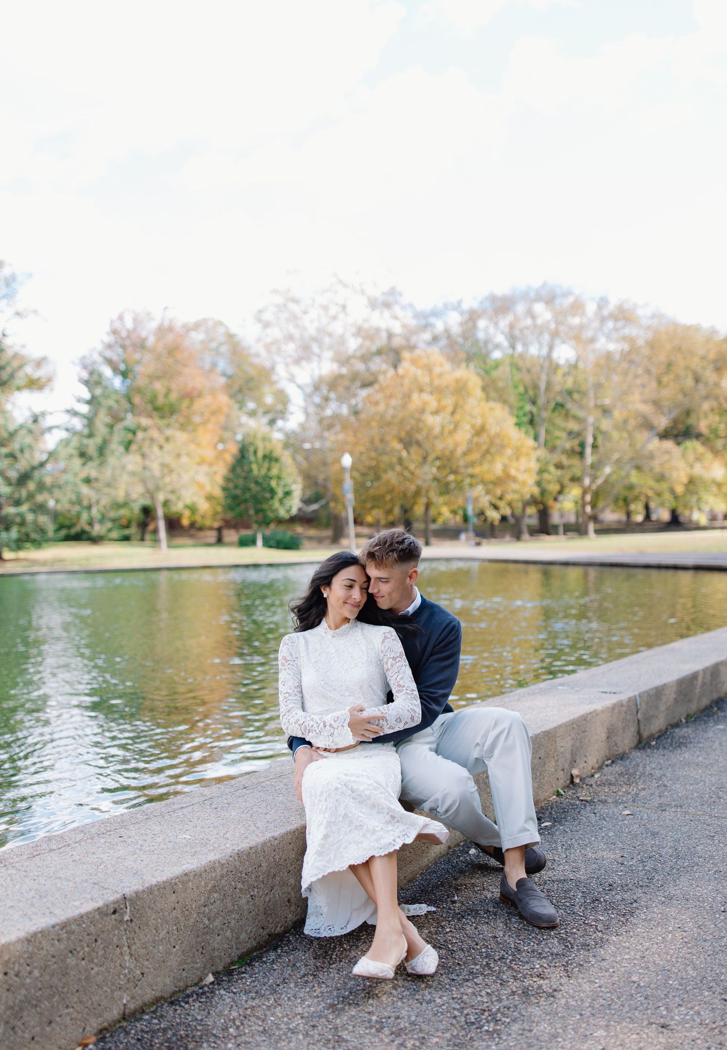 A couple sits together on a concrete wall by a serene lake during autumn with trees reflecting in the water.