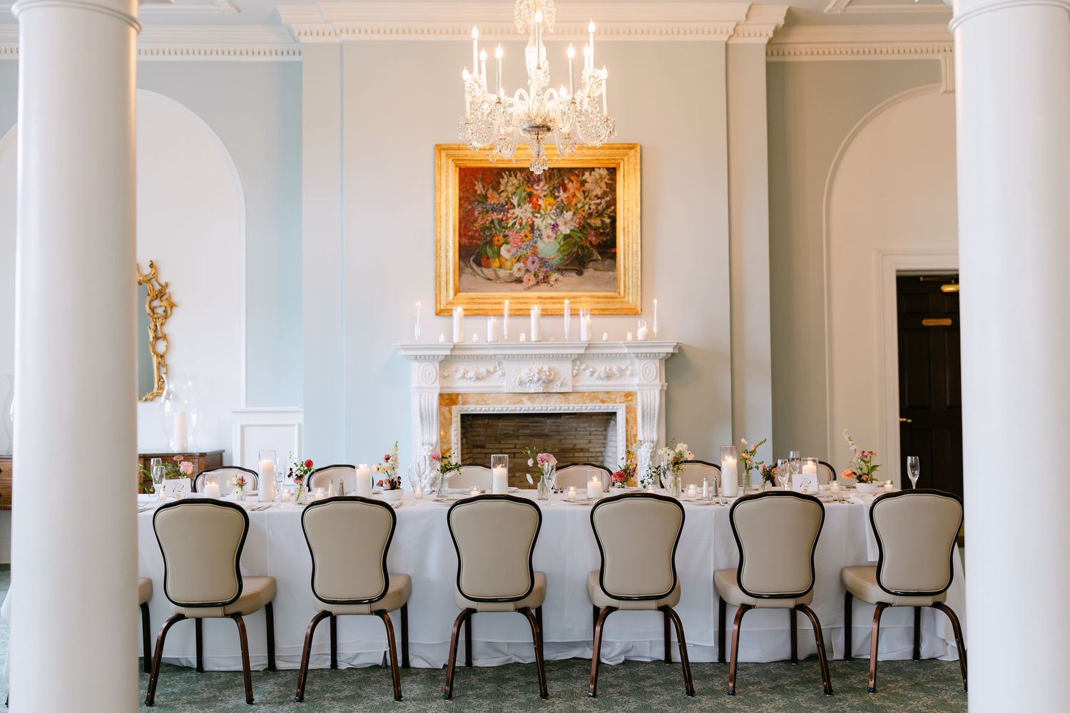 Elegant white dining room with crystal chandelier, ornate fireplace, and row of upholstered chairs at long table.