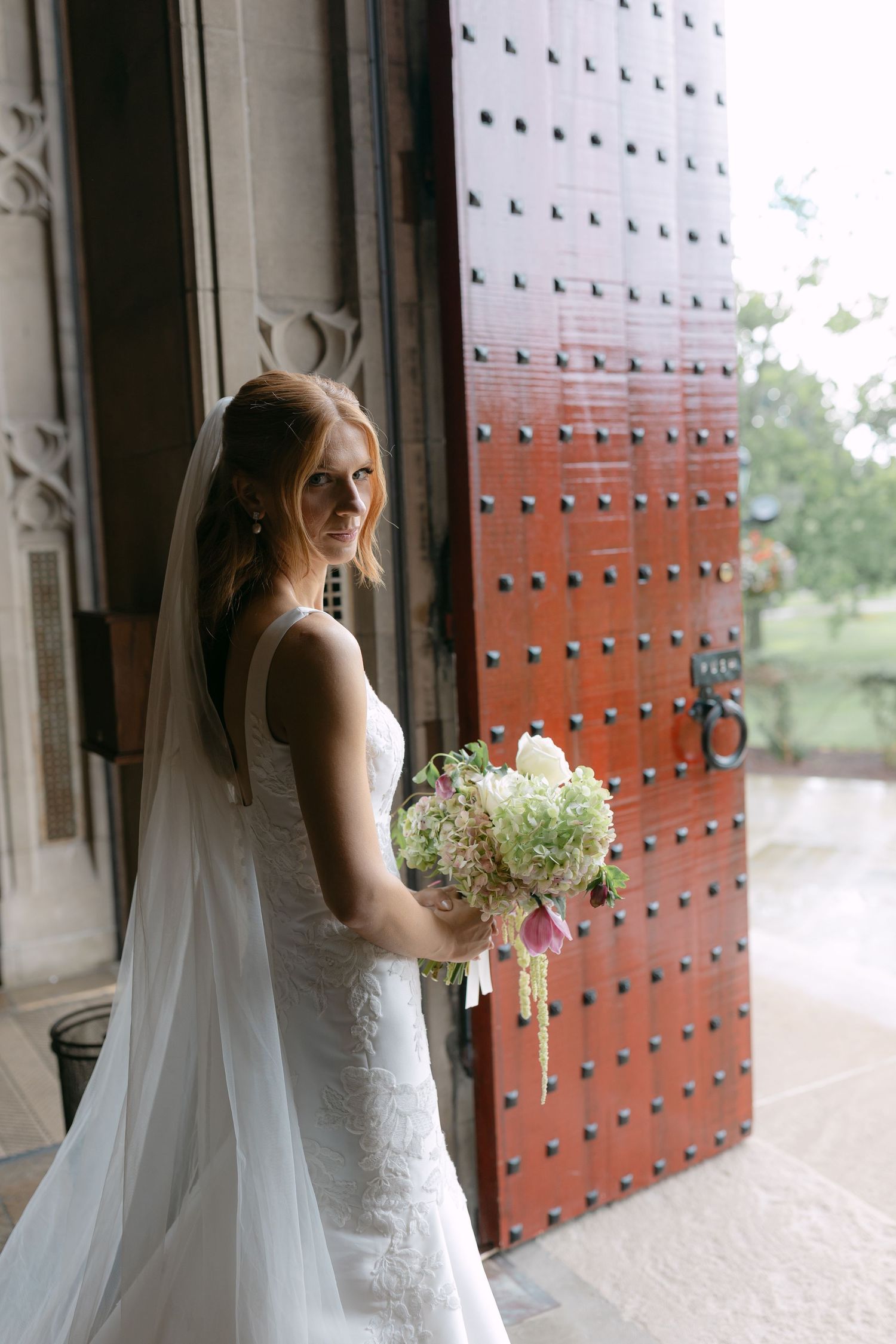 Bride in white lace gown holding floral bouquet stands before ornate red door with veil flowing.