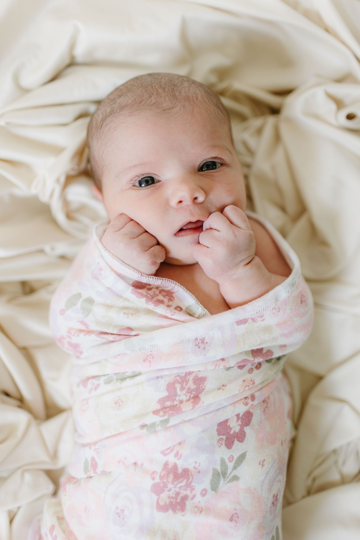 Newborn baby wrapped in floral swaddle blanket lying on cream colored fabric.