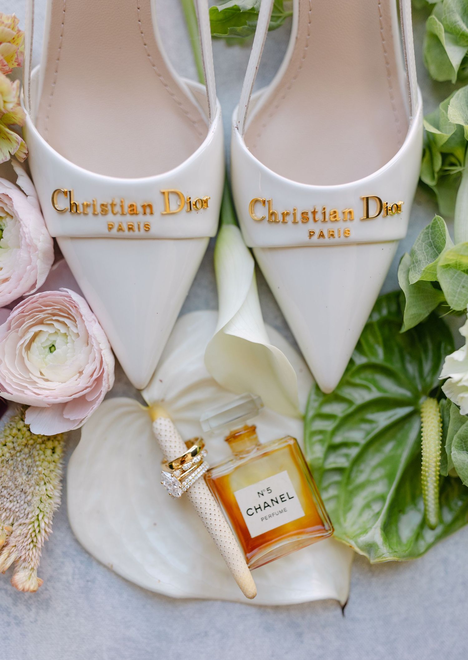 Elegant white Christian Dior bridal shoes displayed with perfume bottles and delicate pink flowers against a light background.
