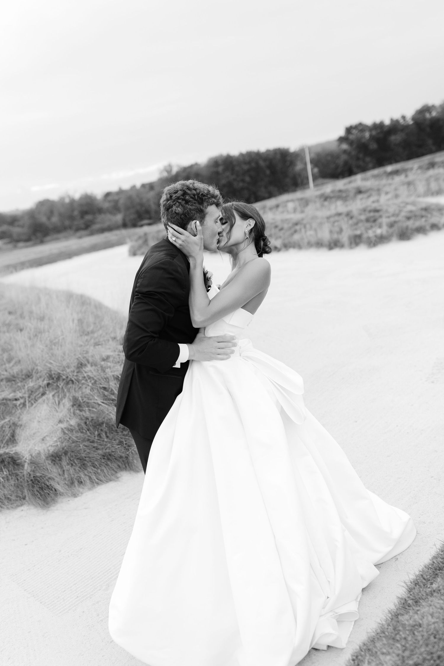 Black and white photo of a bride and groom sharing a romantic kiss outdoors on their wedding day.