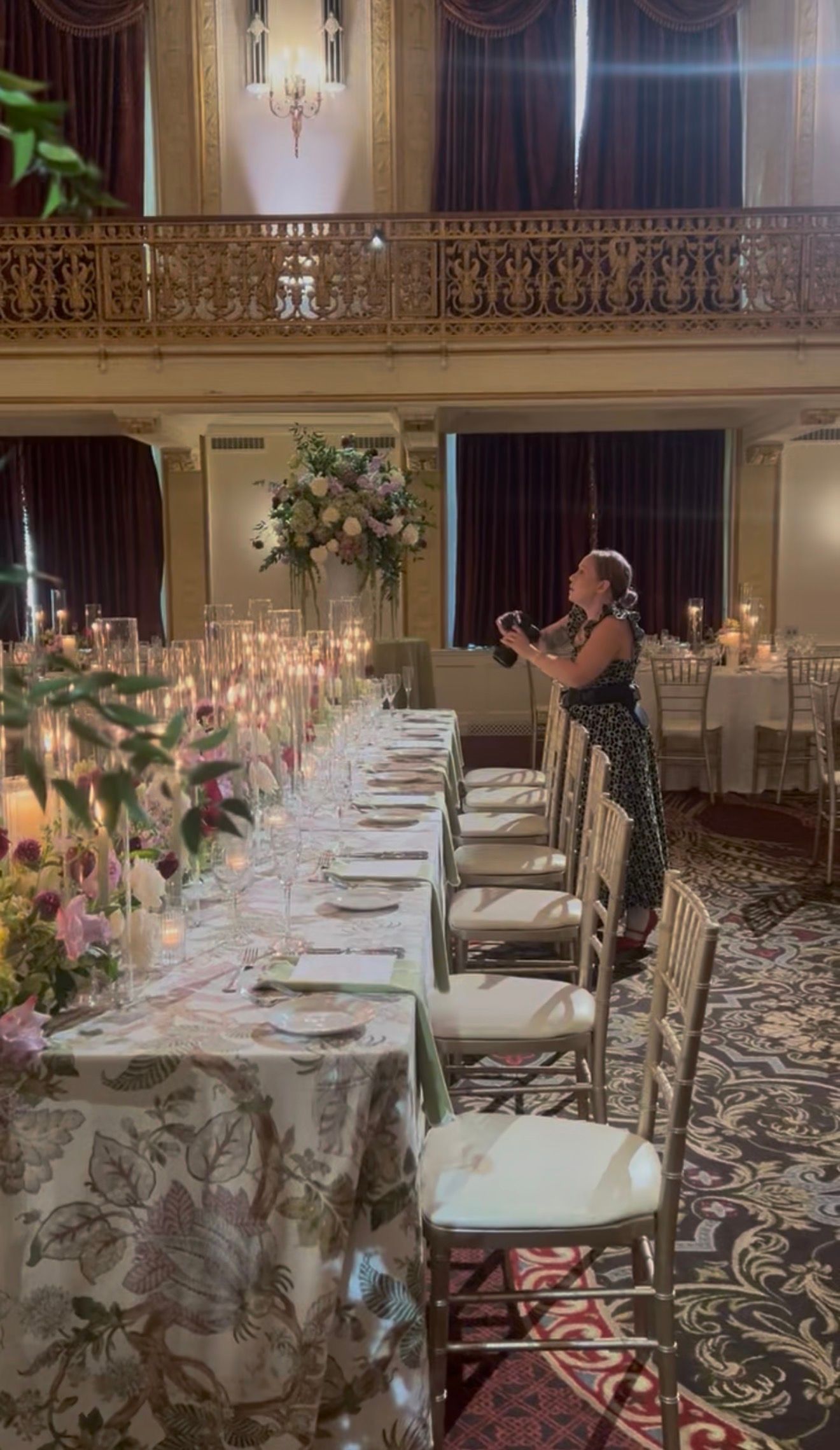 Elegant banquet table settings with white linens, floral arrangements and glowing candles in an ornate ballroom with patterned carpet.