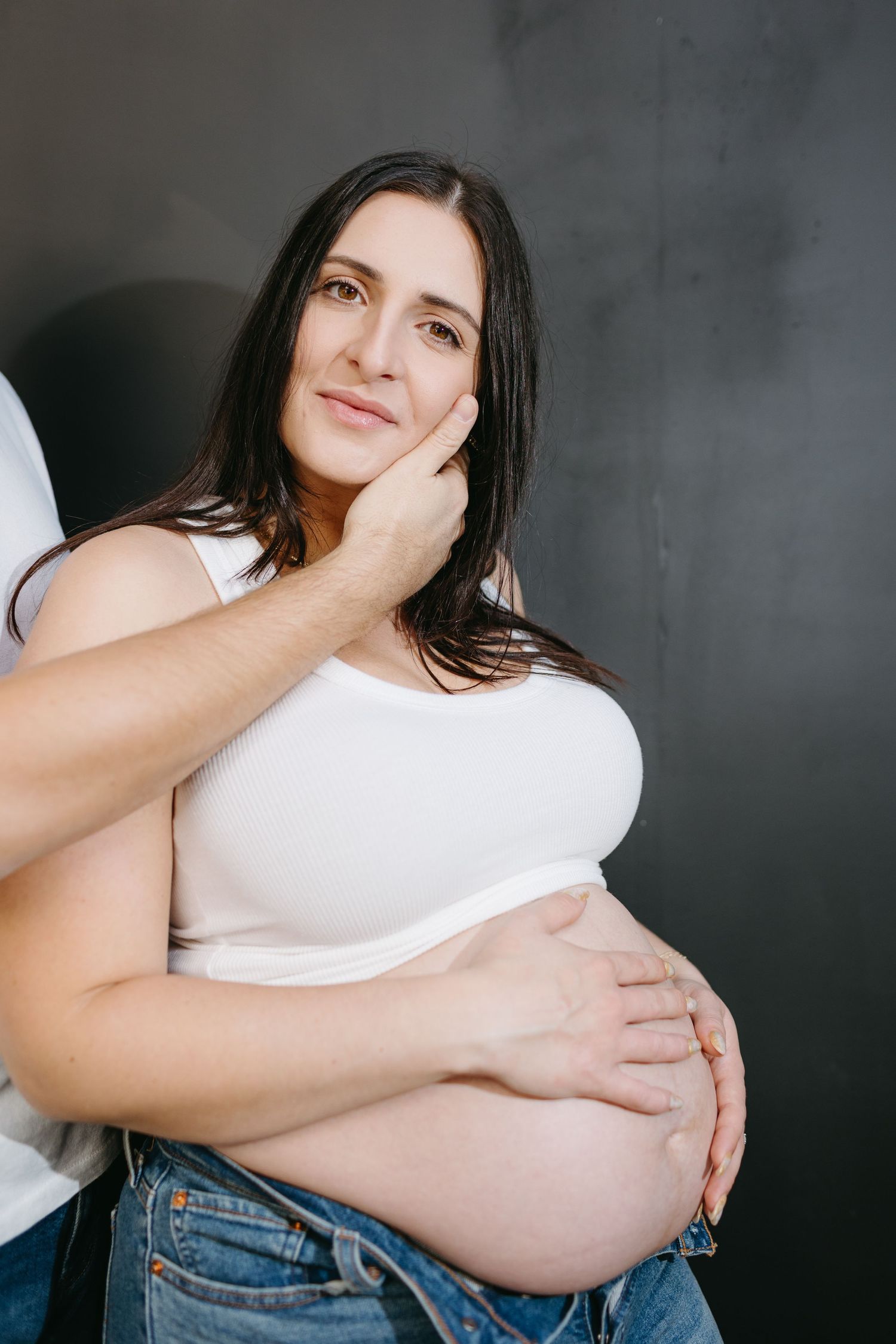 Side view of pregnant woman in white top touching her belly against dark background.