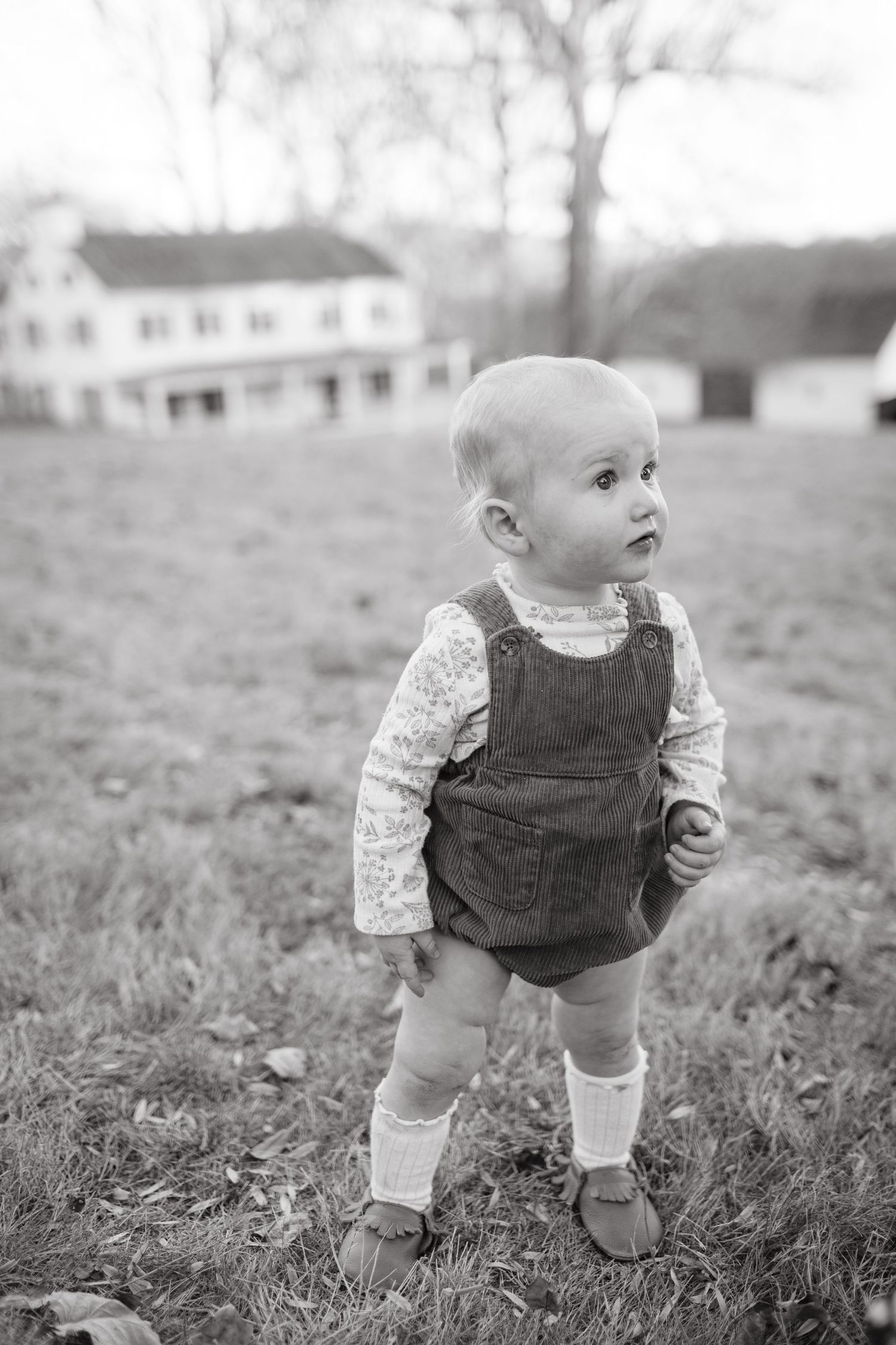 A small child in overalls and knit sweater stands in a field with a farmhouse in the background.