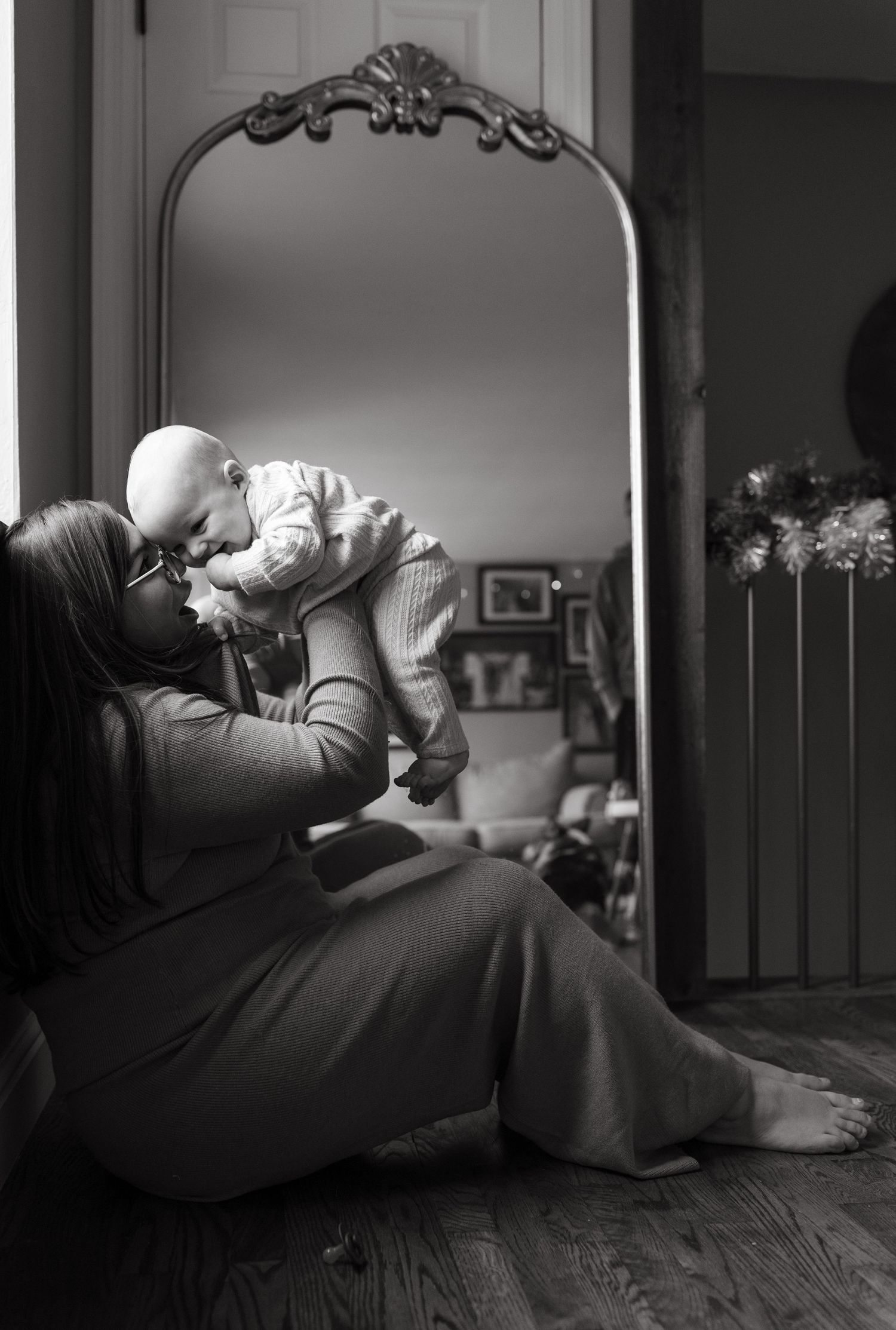 Intimate black and white portrait showing tender moment between parent and baby in front of ornate mirror.
