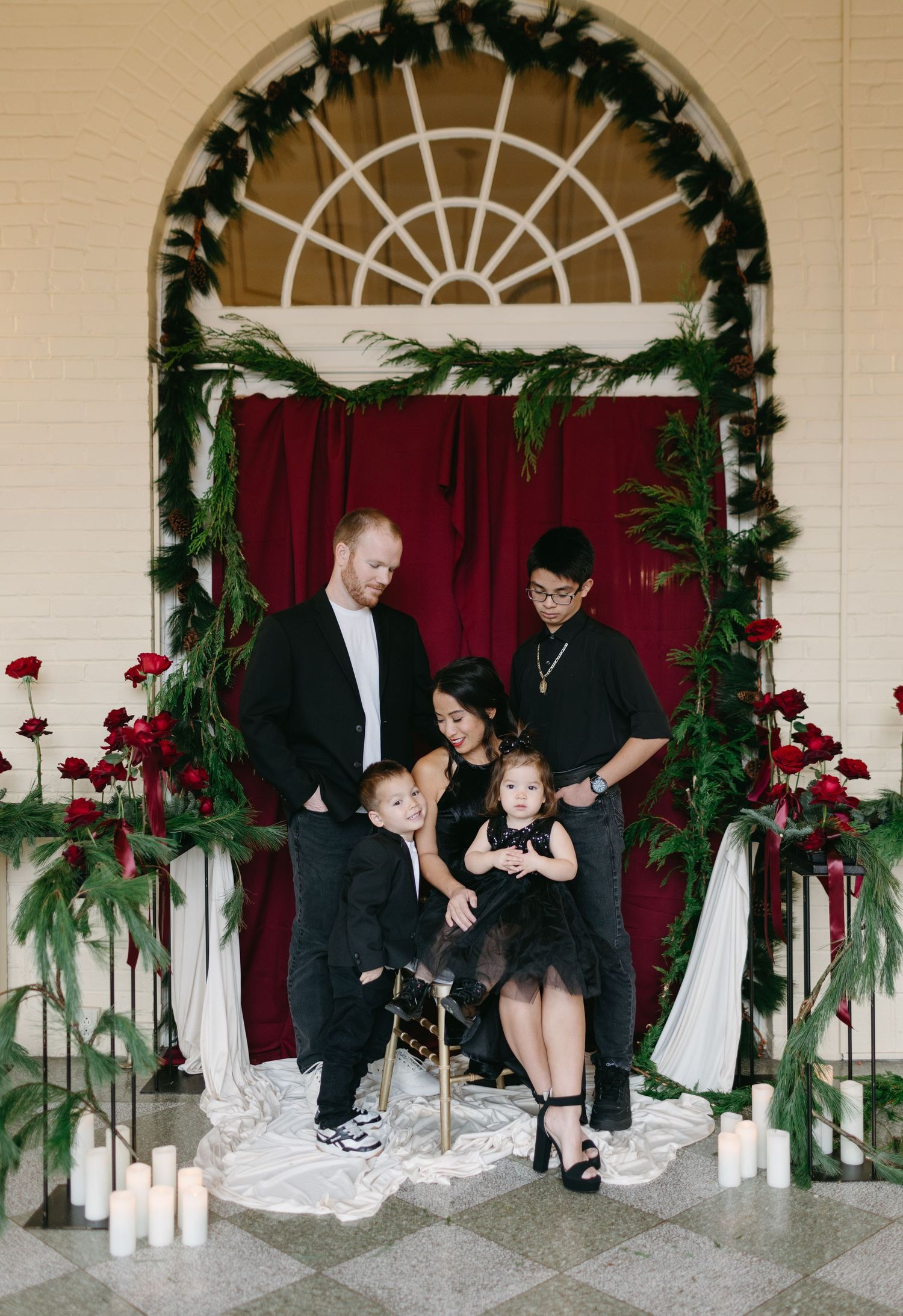 Family portrait in formal black attire against red curtain with holiday greenery and candle decorations.