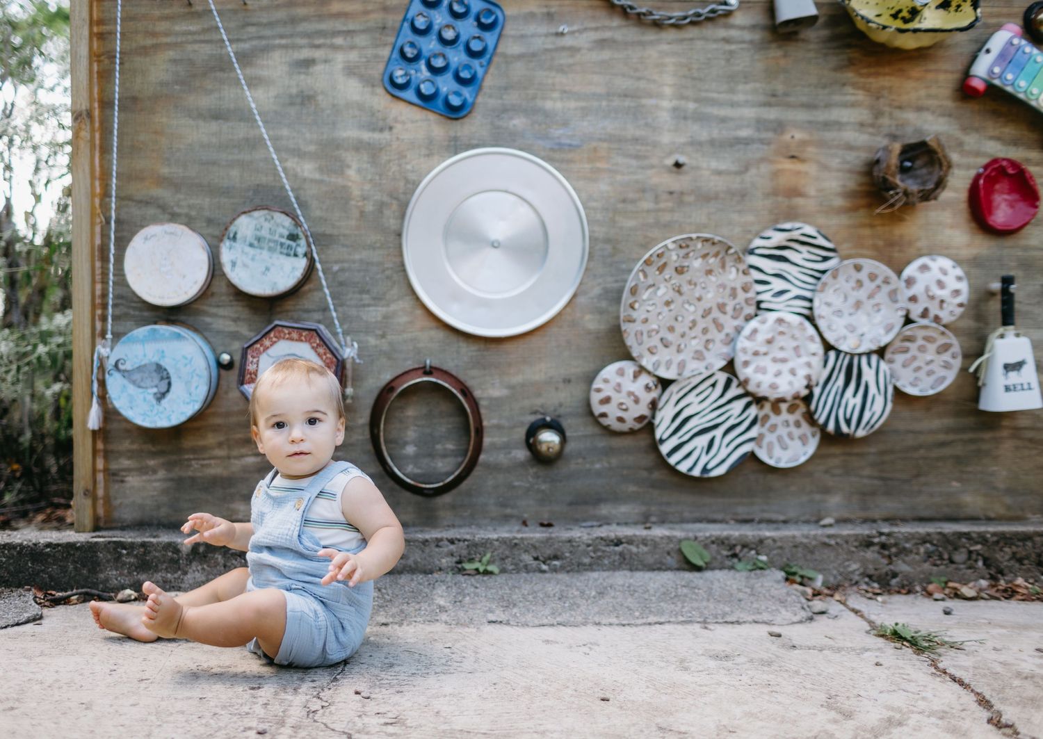 Baby in striped outfit sitting by vintage wall display with decorative plates and objects.