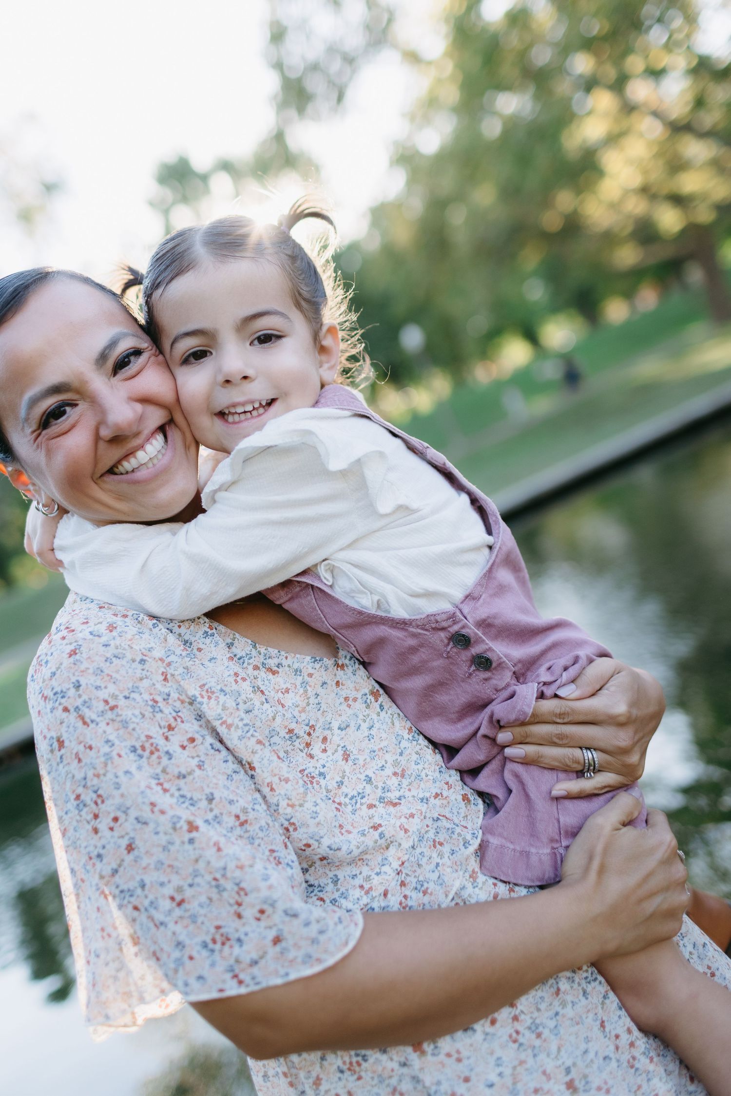 A tender moment of joy and laughter shared outdoors on a sunny day near a pond.