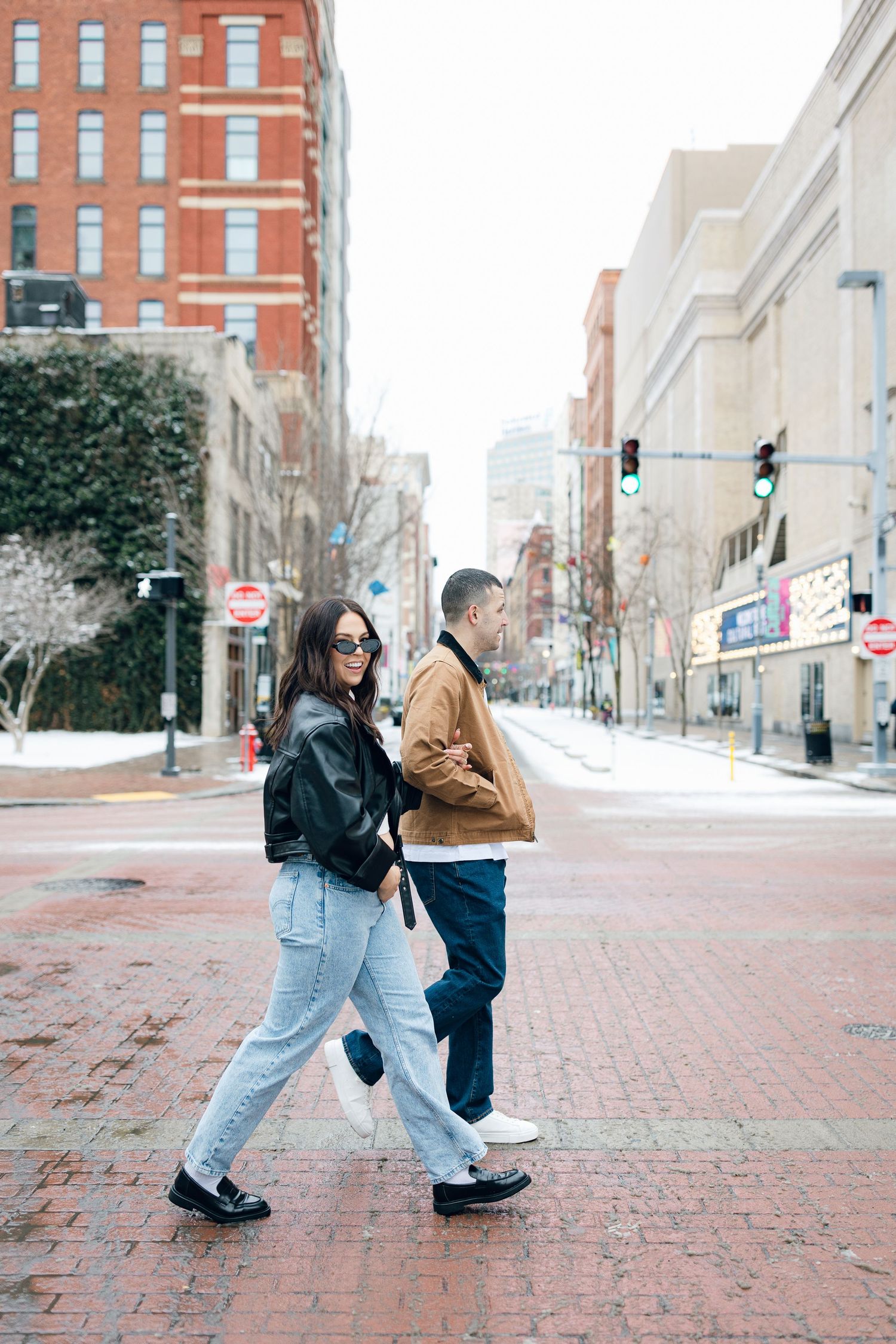 A stylish couple walking down a snowy urban street with brick buildings in the background.