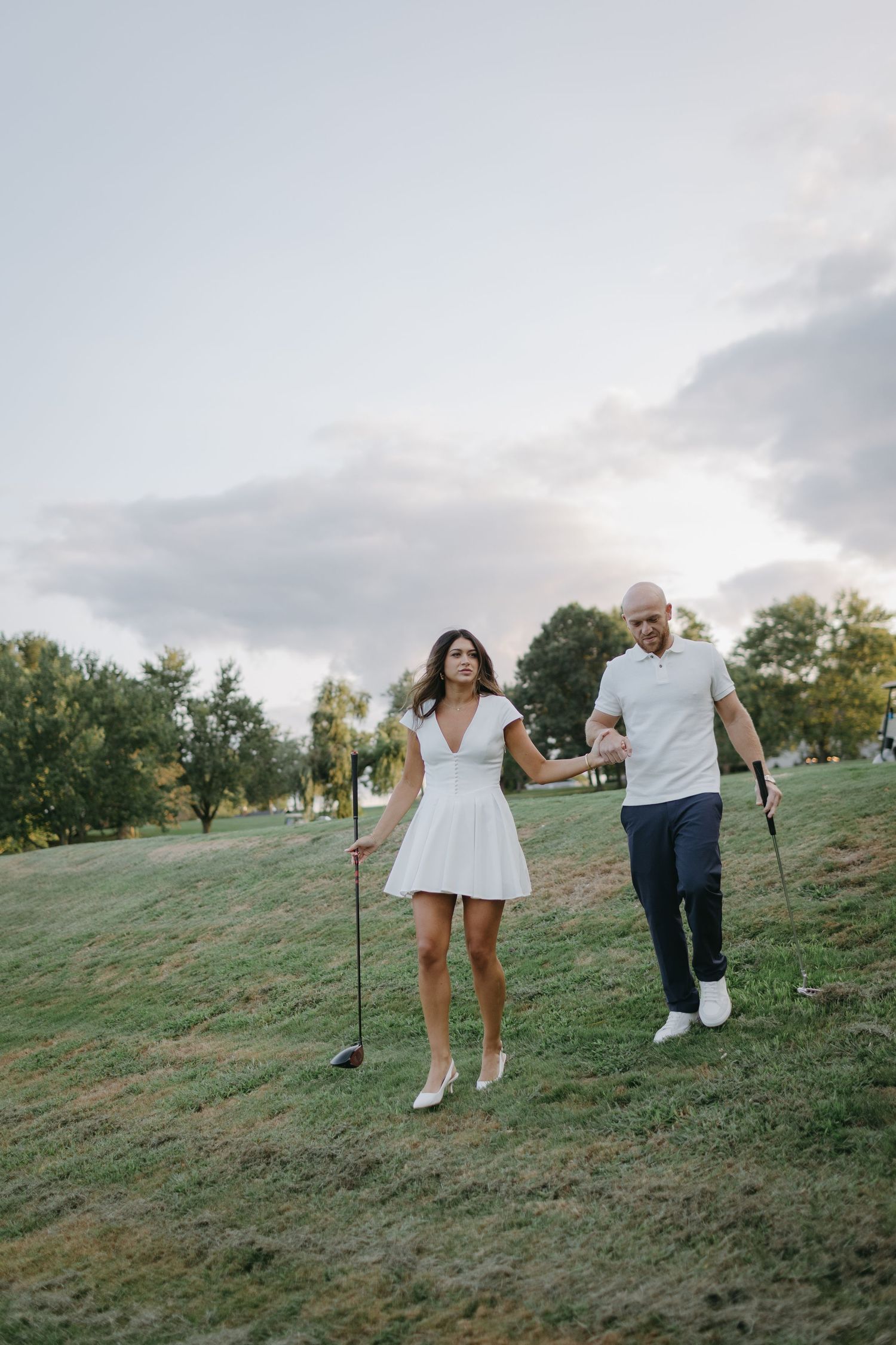Couple holding golf clubs while walking uphill on golf course during sunset.