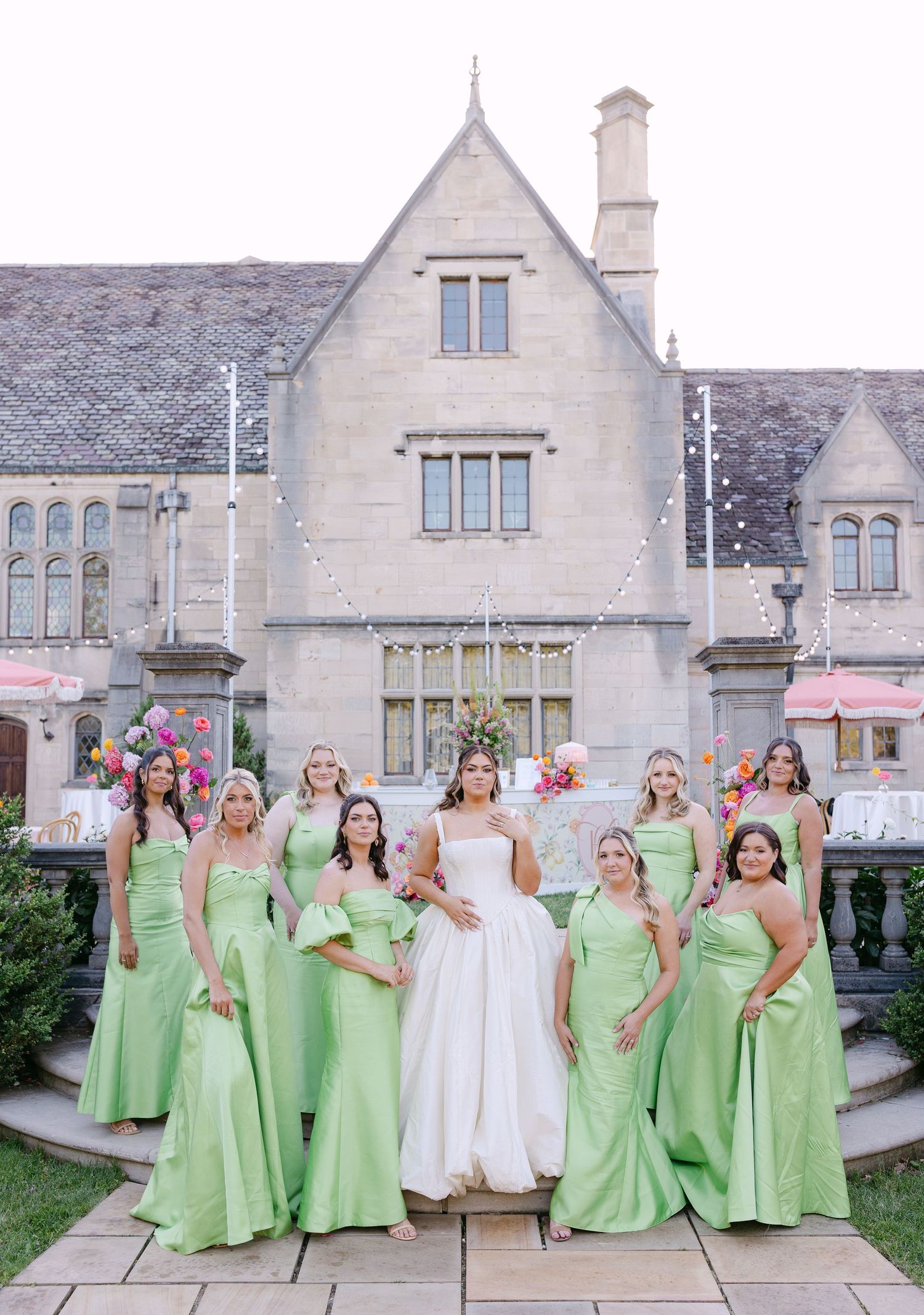 Bride in white gown poses with eight bridesmaids in mint green dresses outside an elegant stone manor.