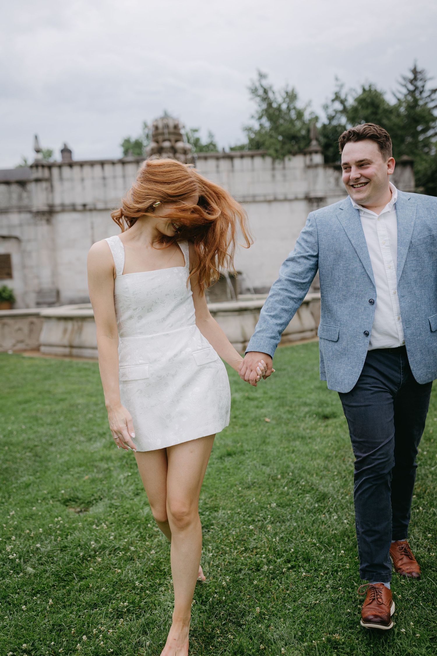 A couple playfully runs through a grassy field, with the person in white spinning around while holding hands.